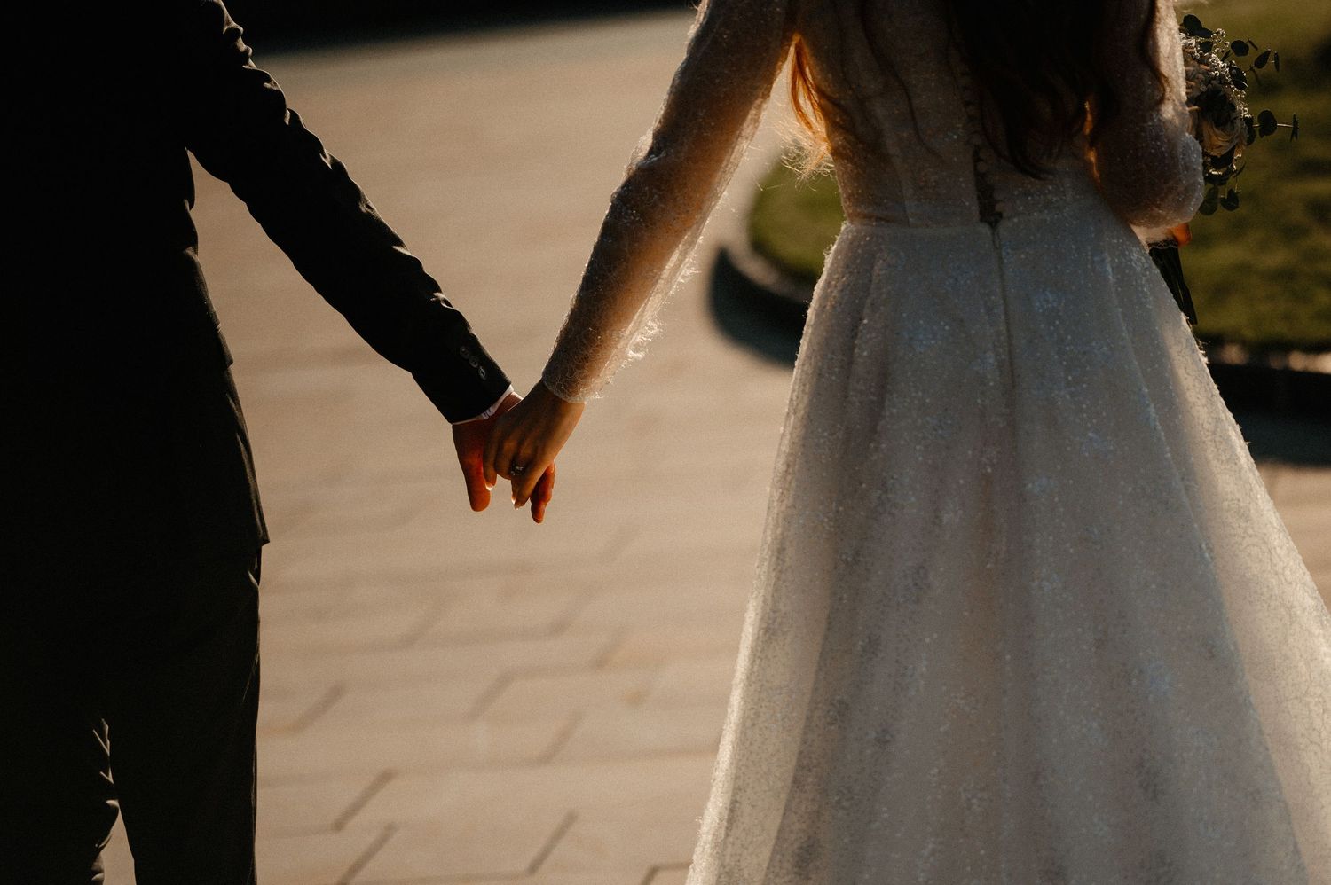 Bride and groom holding hands walking on stone path in dramatic shadowed lighting.