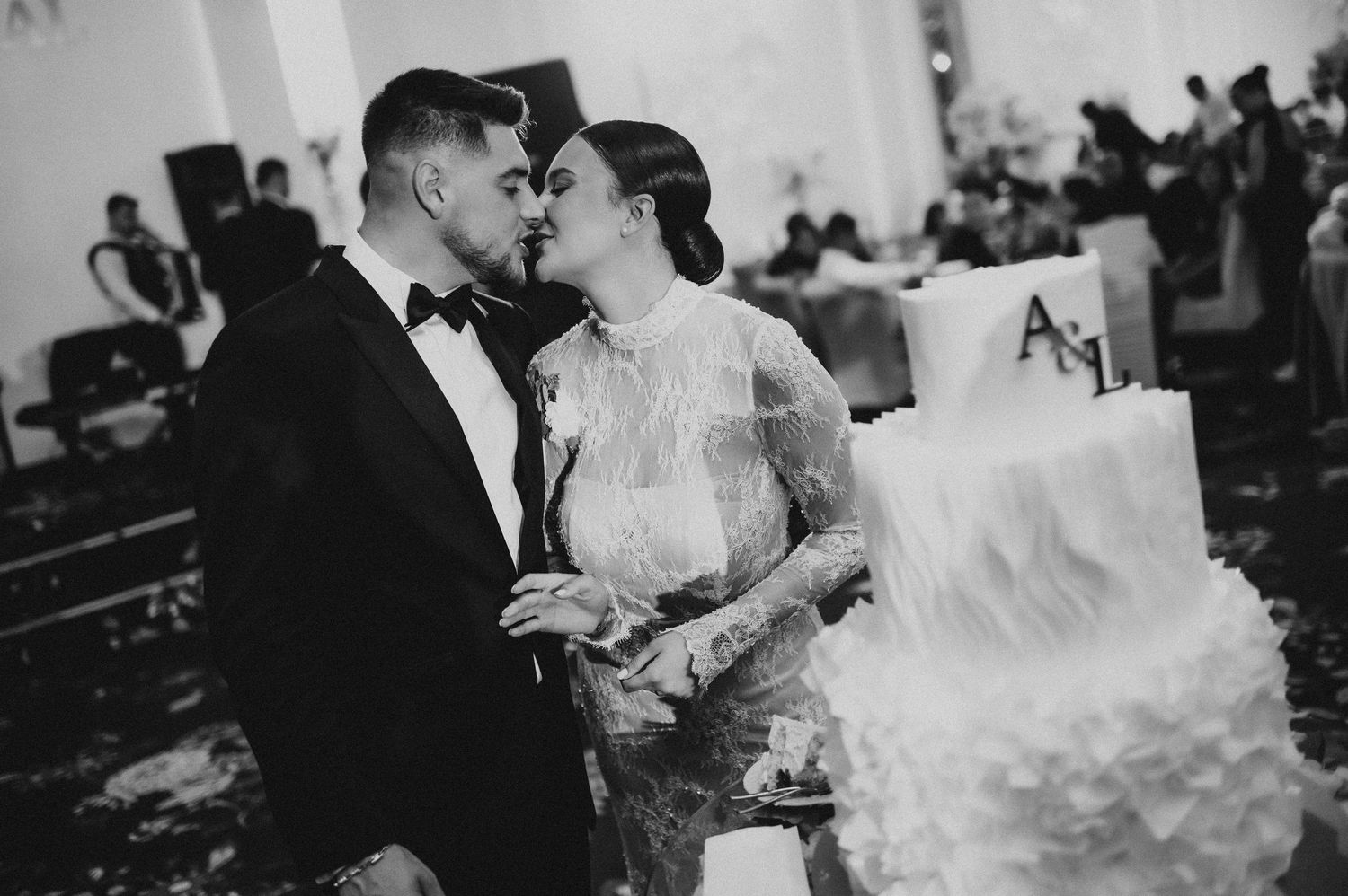 Black and white photo of newlyweds kissing beside their elegant white ruffled wedding cake.