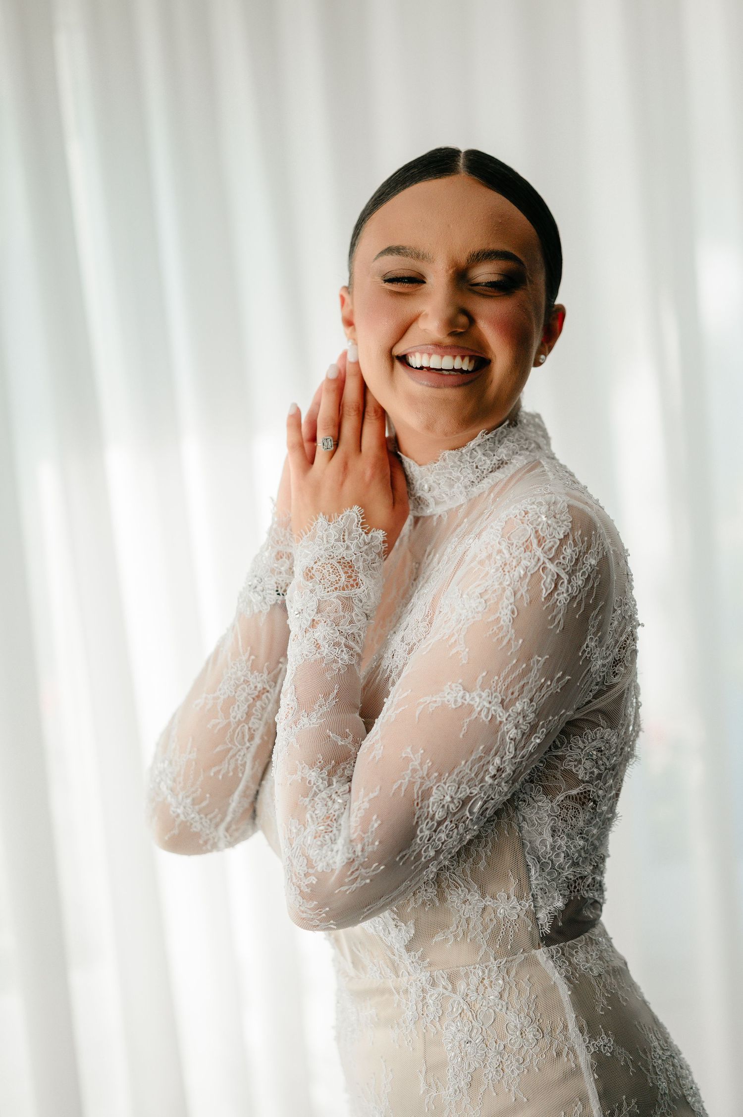 Joyful bride in a high-neck long-sleeve lace wedding dress laughing with hands raised to her face.