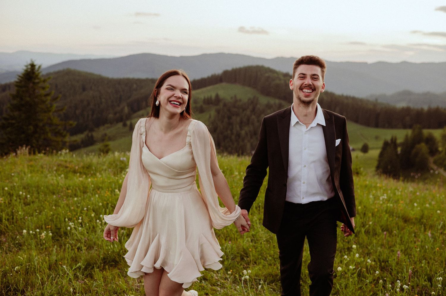 Happy bride and groom holding hands and laughing in a green mountain meadow.