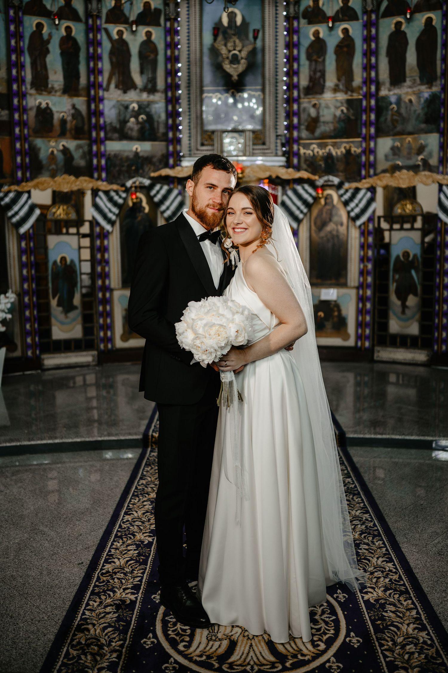 Bride and groom posing together inside Orthodox church with ornate iconostasis backdrop.