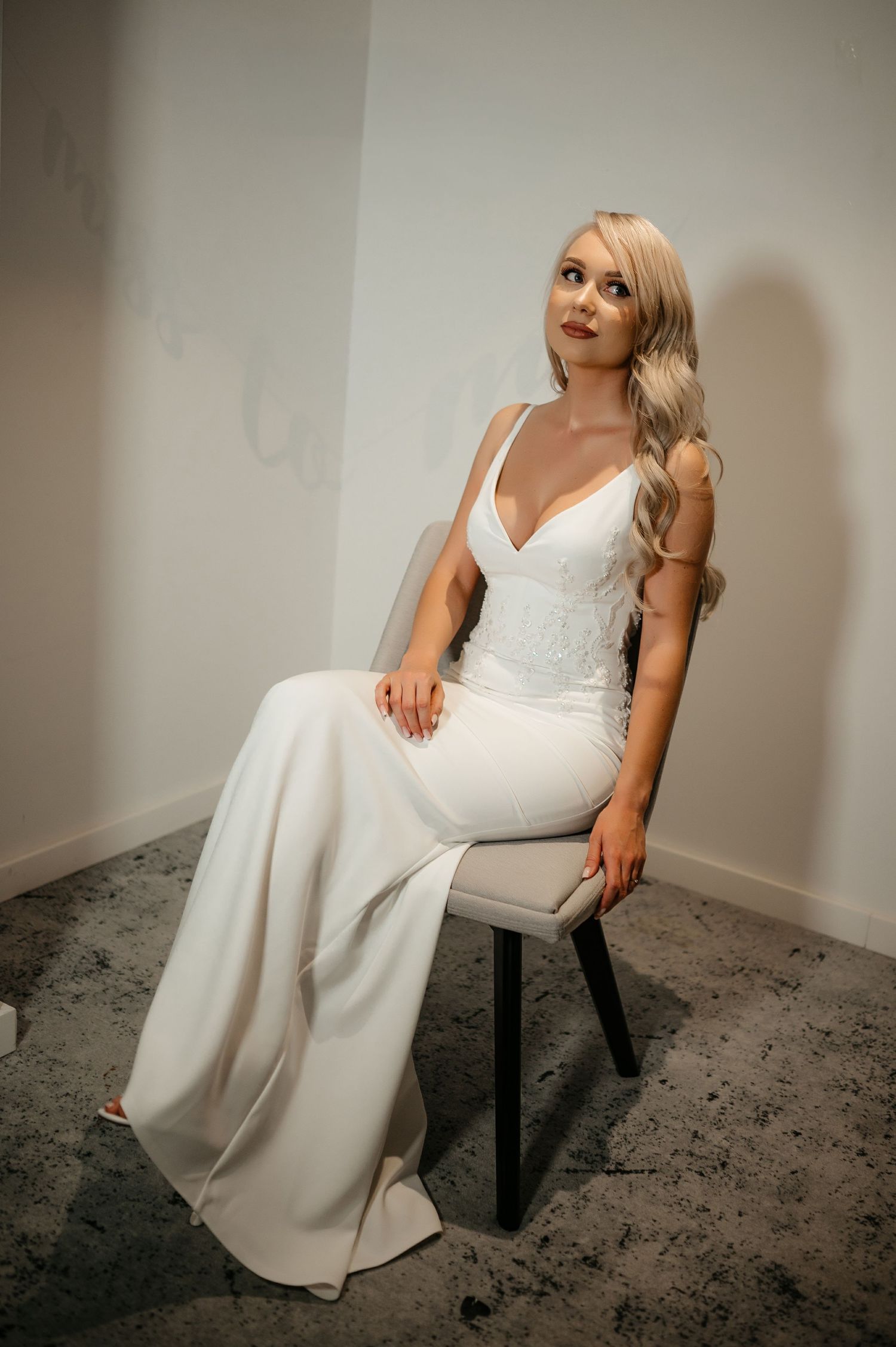 Blonde bride in a sleeveless white wedding dress with lace bodice seated on a chair looking upward.