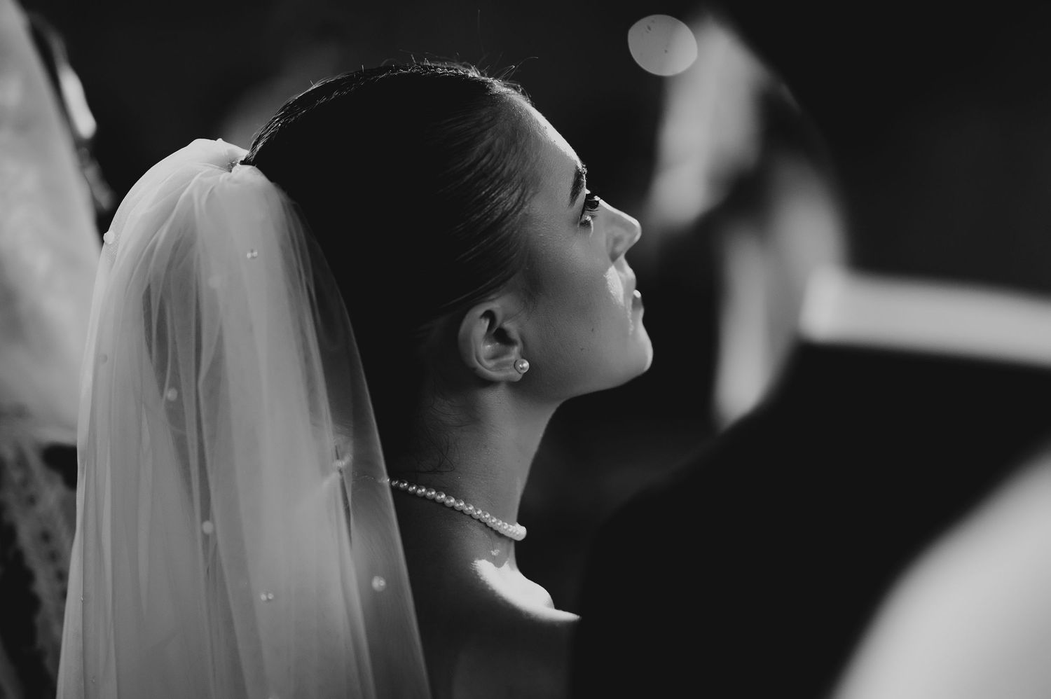 Elegant black and white profile portrait of bride wearing veil and pearl necklace at ceremony.