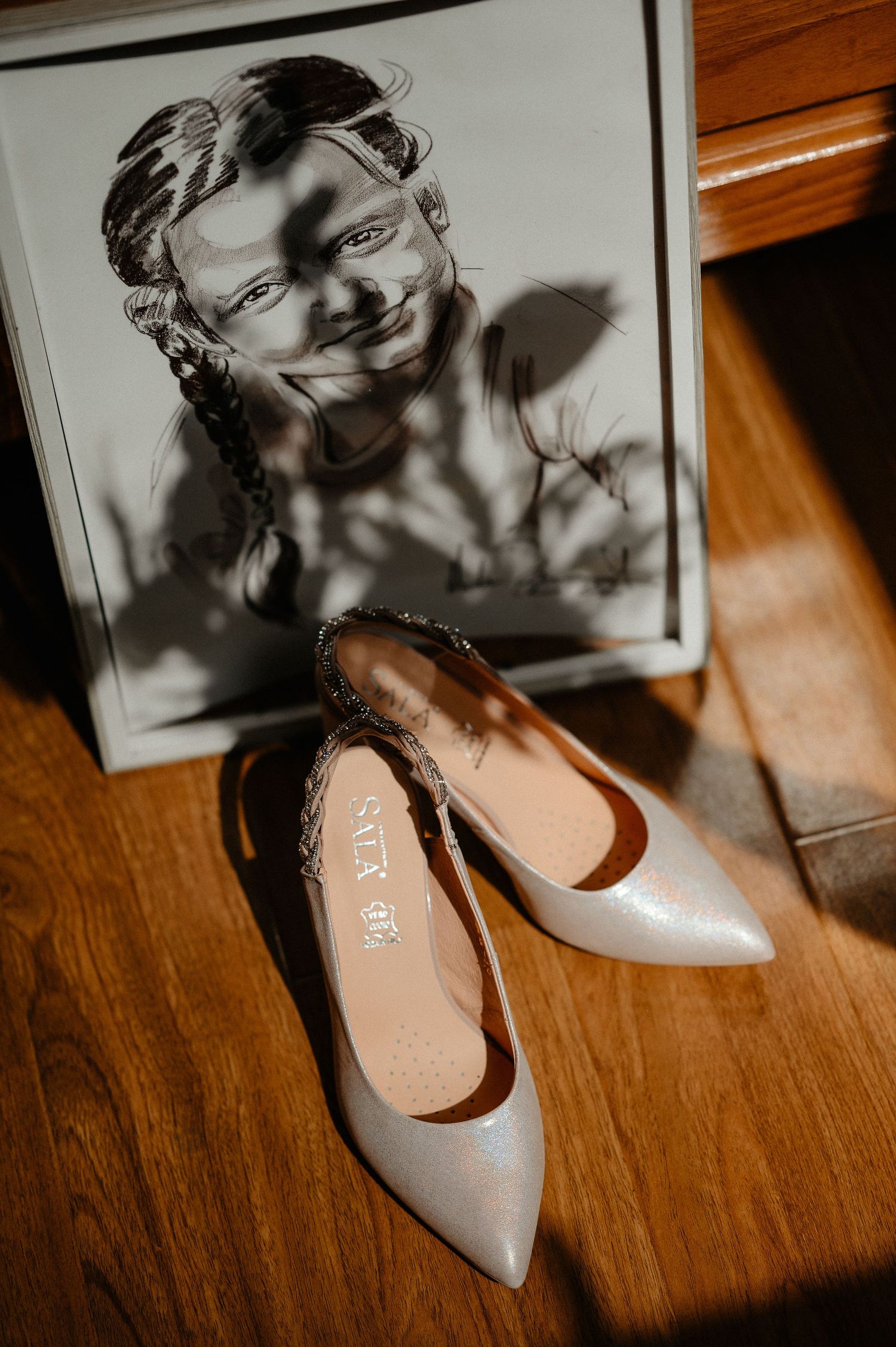 Elegant white bridal heels with feather trim displayed on wooden floor beside a charcoal portrait sketch.
