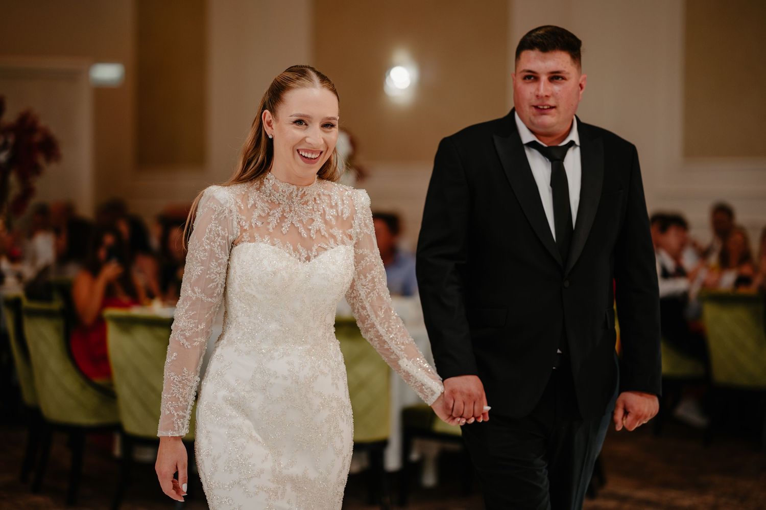 Smiling bride in lace high-neck wedding gown walking hand in hand with groom in tuxedo.