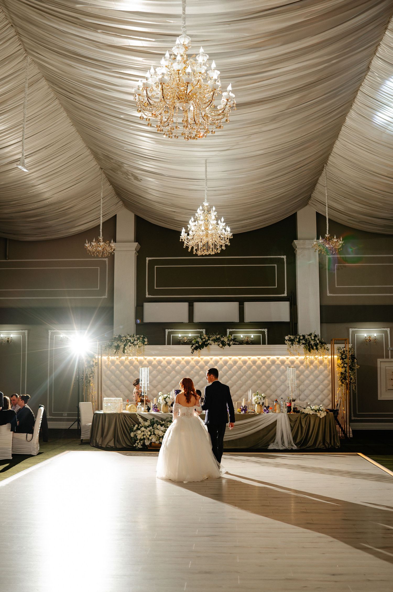 Bride and groom share first dance in elegant ballroom with crystal chandeliers and draped ceiling at wedding reception.