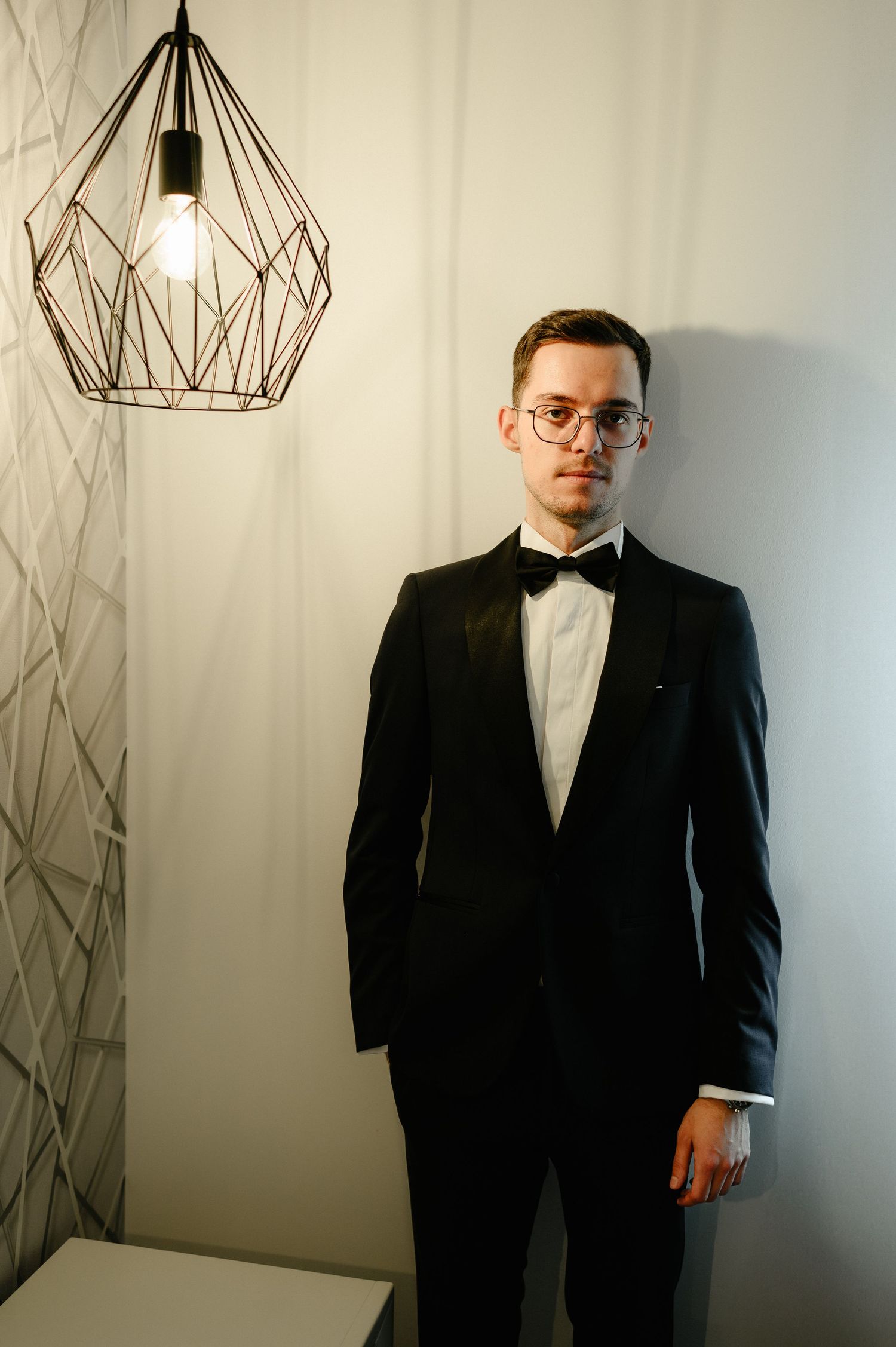 A young man in a black tuxedo and bow tie with glasses stands by a white wall near a geometric pendant light.