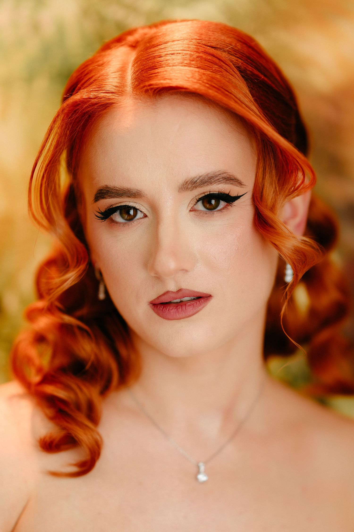 A woman with vibrant red curly hair and elegant makeup wearing a diamond necklace and earrings poses gracefully.
