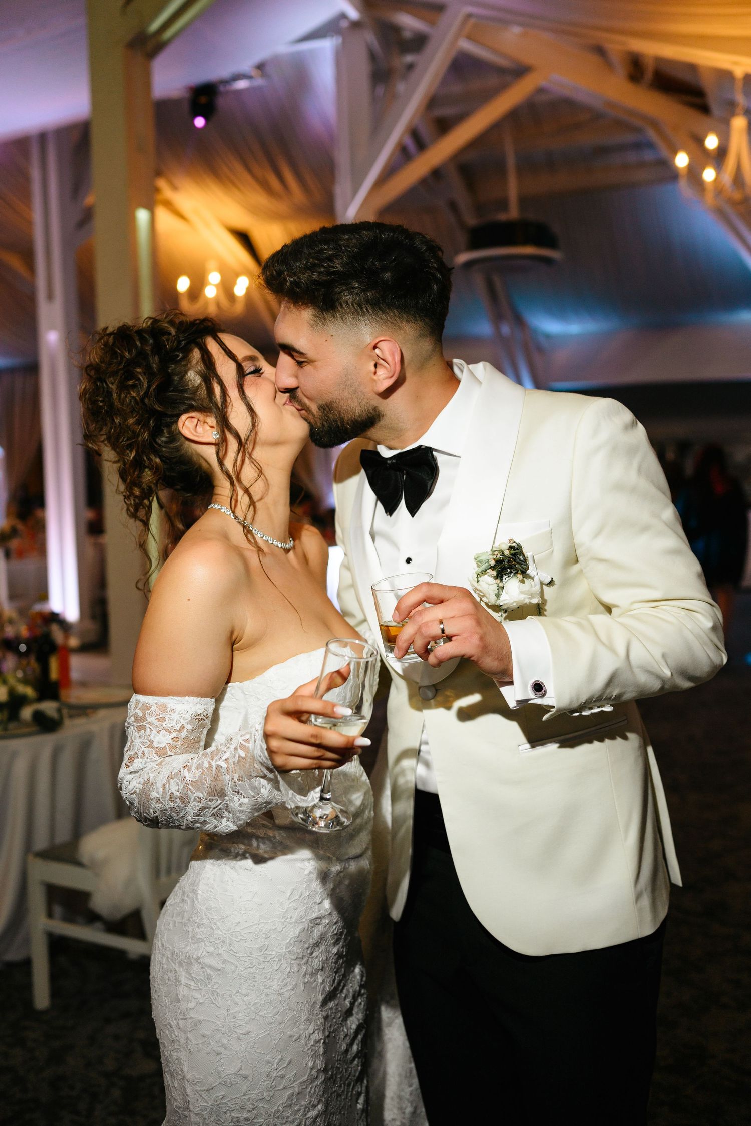 Bride and groom share a romantic kiss while holding champagne glasses at their elegant wedding reception.