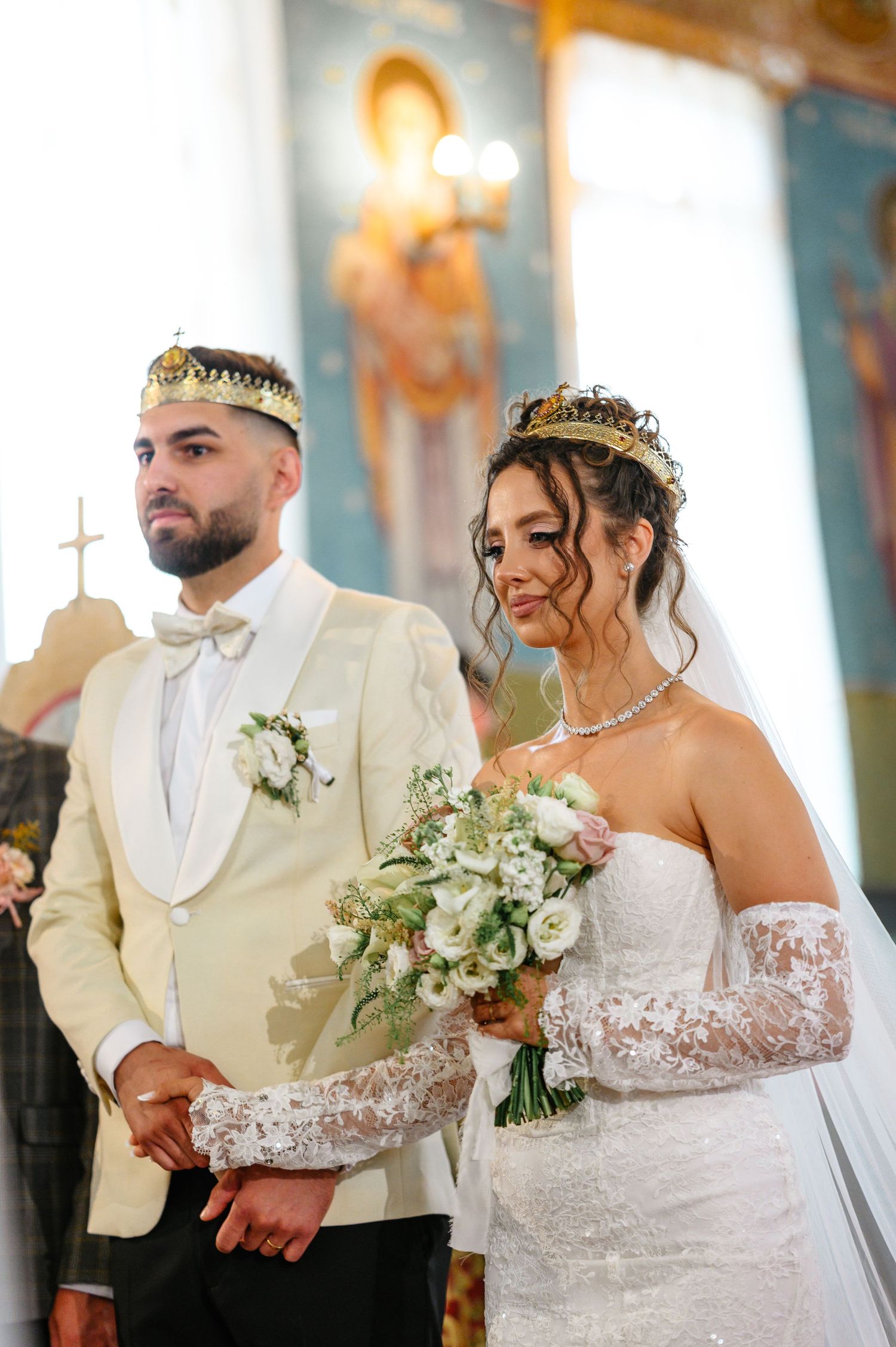 Bride and groom in crowns during Orthodox wedding ceremony, bride holding white floral bouquet in lace gown.
