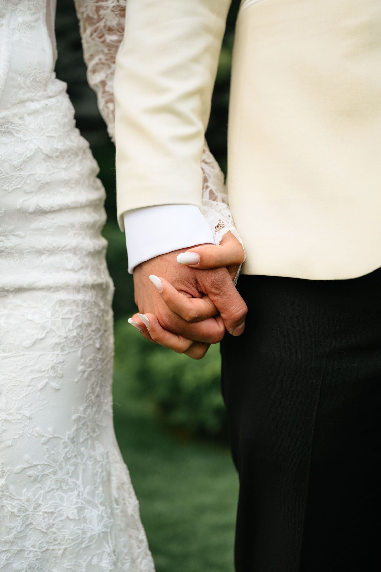 Close-up of bride and groom holding hands, bride in lace dress, groom in cream suit with black pants.