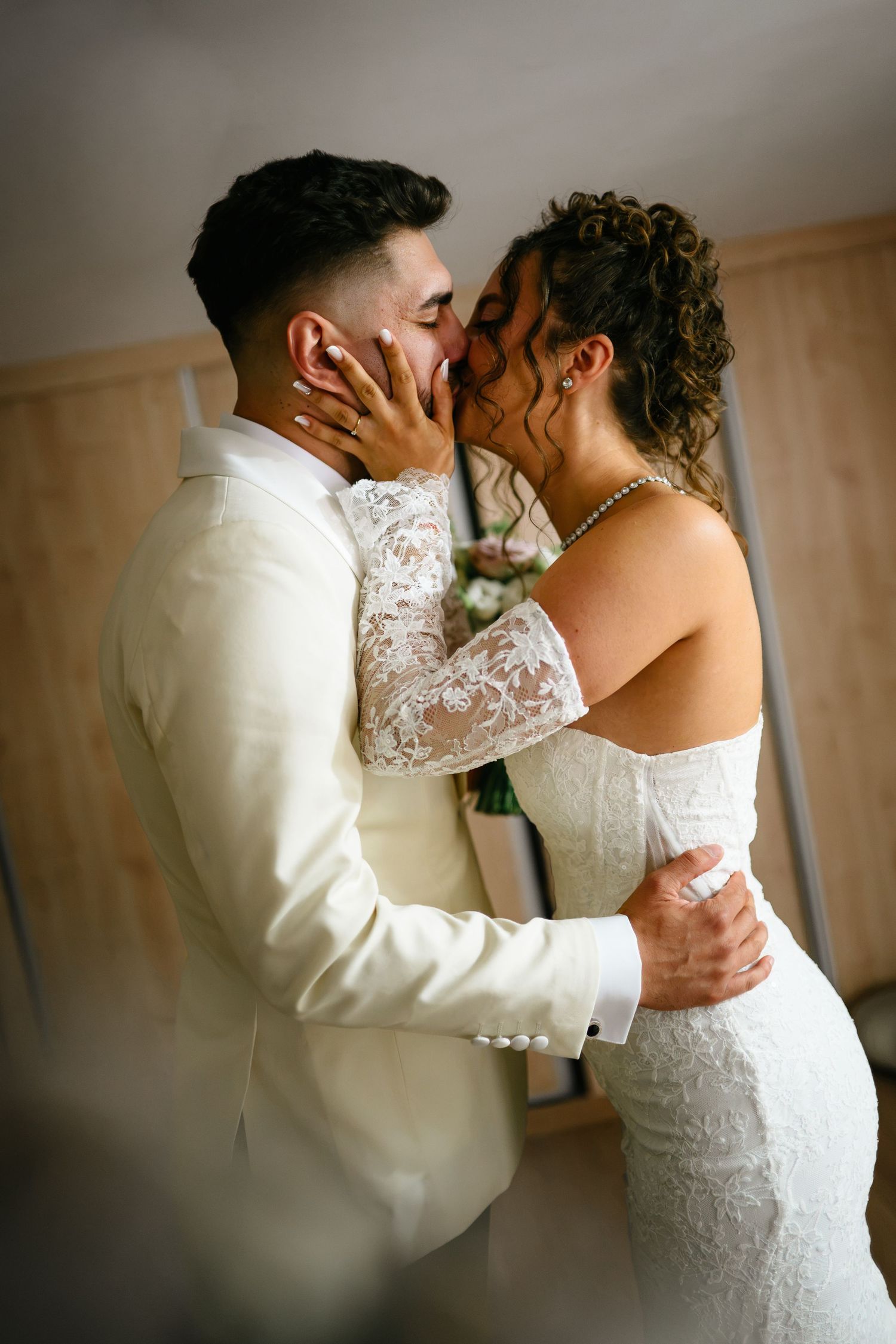 A bride and groom share a romantic first kiss, dressed in elegant white wedding attire in a softly lit room.