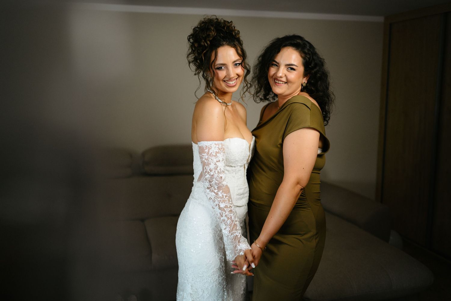 Bride in white lace gown poses with woman in olive dress, both smiling and holding hands indoors.