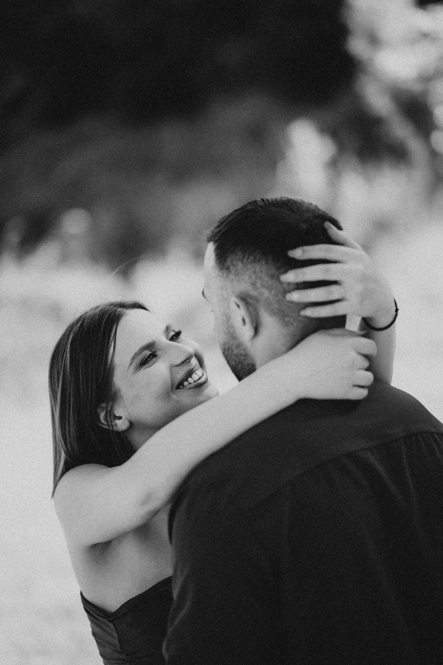 Black and white photo of laughing woman embracing man outdoors in romantic moment.