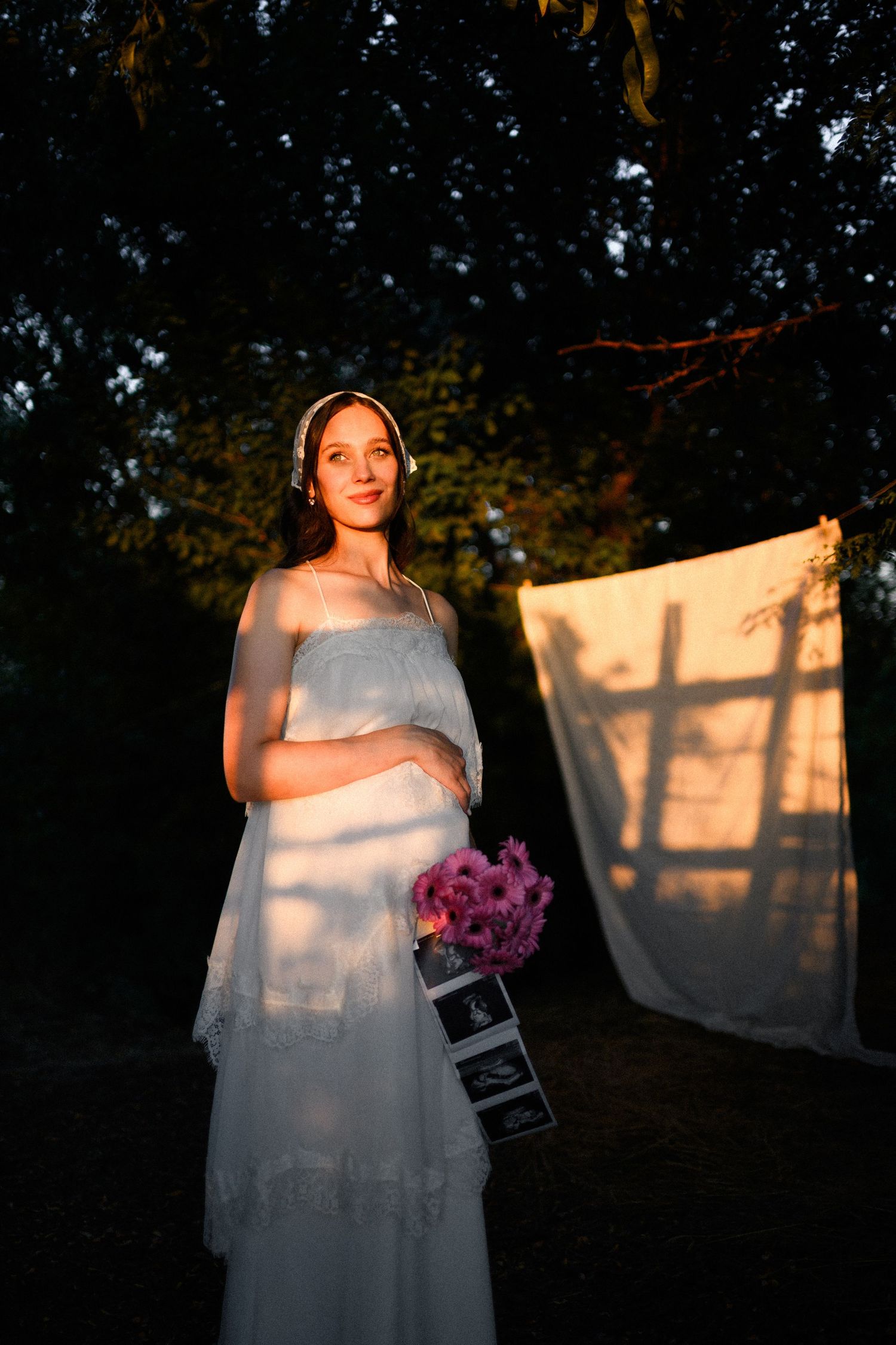 Pregnant woman in white dress holding flowers and ultrasound photos, bathed in golden sunset light outdoors.