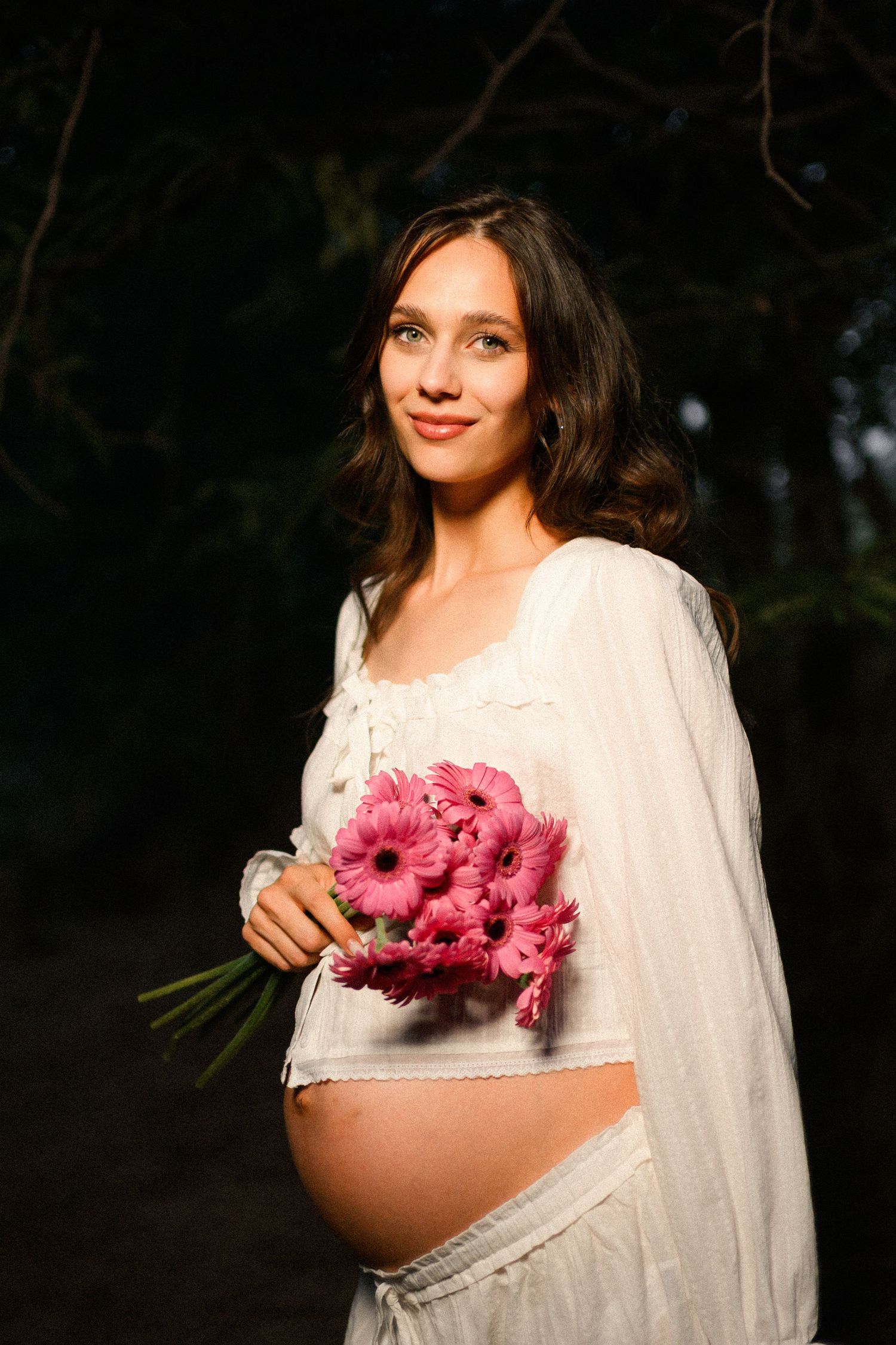 Pregnant woman in white dress holding pink gerbera daisy bouquet, smiling outdoors in natural sunlight.