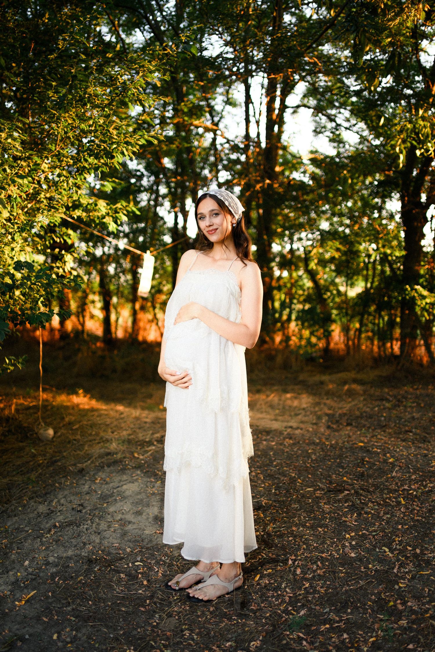 Pregnant woman in white tiered dress and floral headband cradling her baby bump outdoors in golden sunlight.