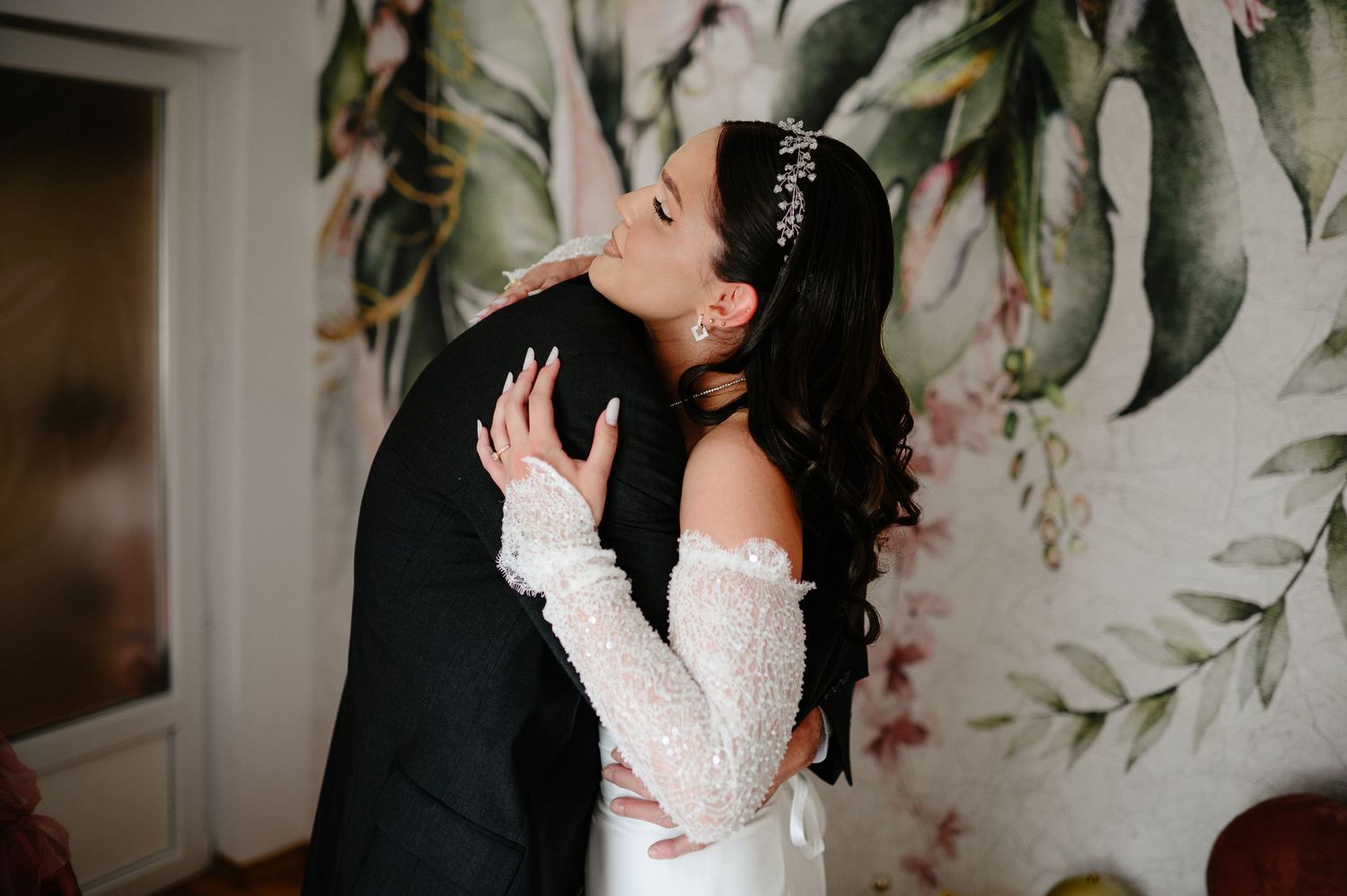 Bride in lace wedding gown sharing romantic kiss with groom in black suit against floral wallpaper.