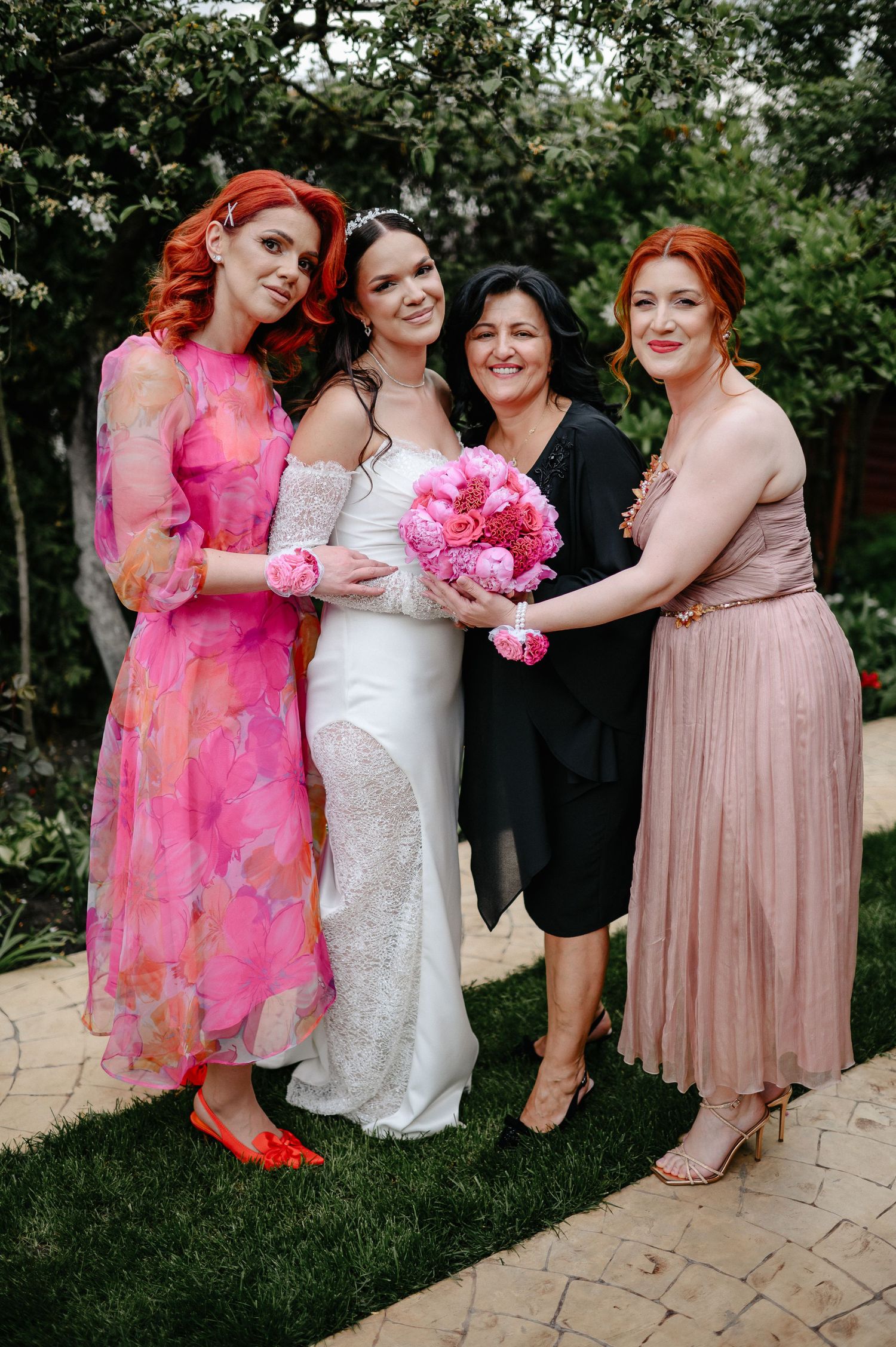 Bride in white gown holds pink peony bouquet surrounded by three women in pink dresses at wedding.