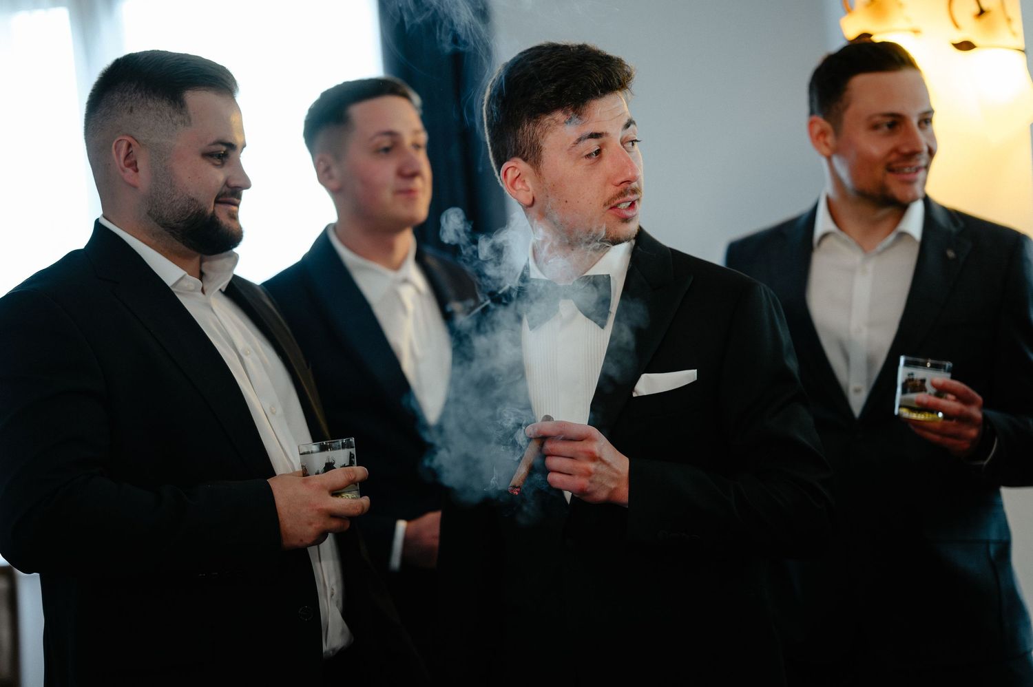 Groomsmen in black suits sharing drinks and cigars before wedding.