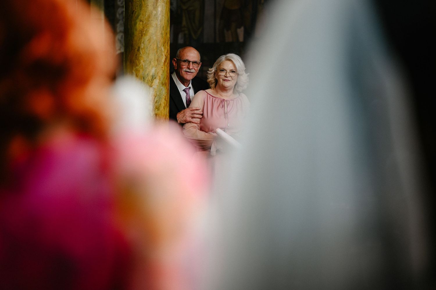 Elderly couple watching a wedding ceremony through a soft-focus foreground of pink flowers and white veil.