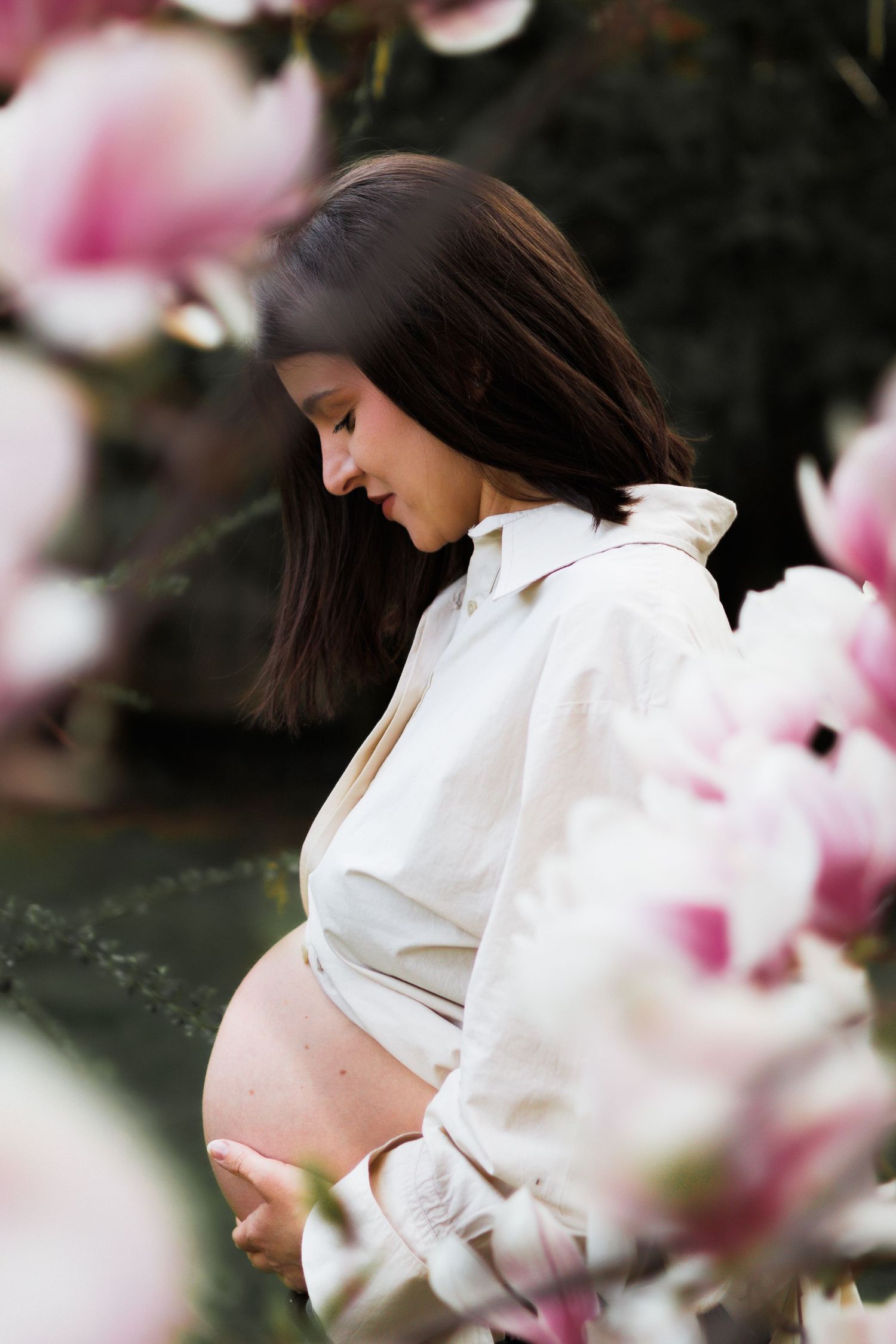 Side profile of pregnant woman in white shirt cradling bump near pink magnolia flowers.