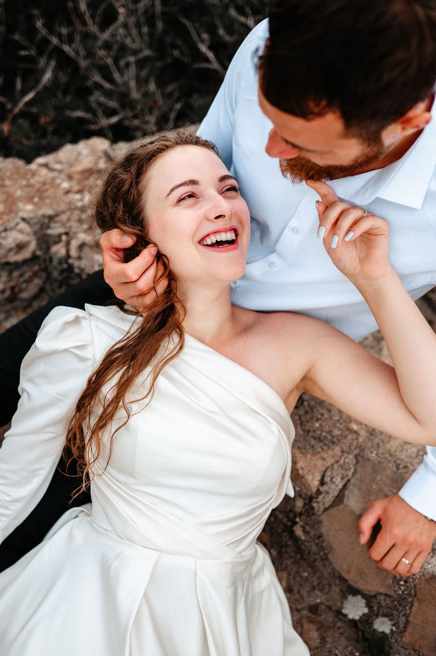 Joyful bride in white satin gown laughs while lying back in groom's arms on rocky terrain.