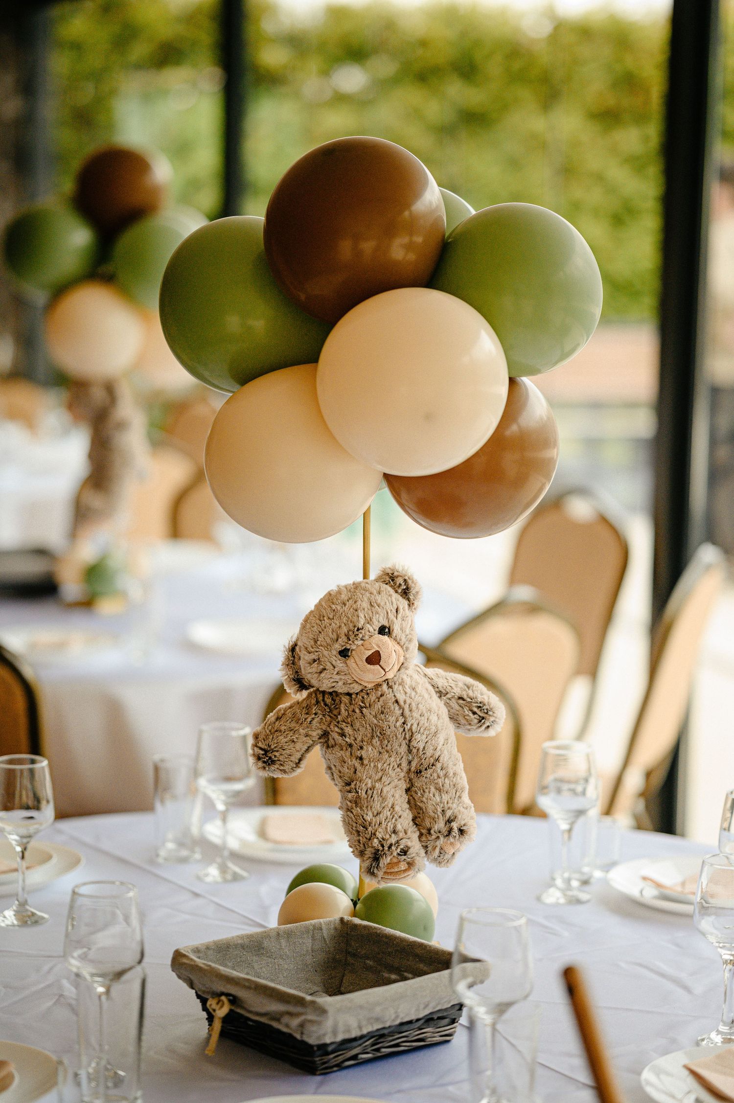 Teddy bear centerpiece with green, brown, and cream balloons on a white table.