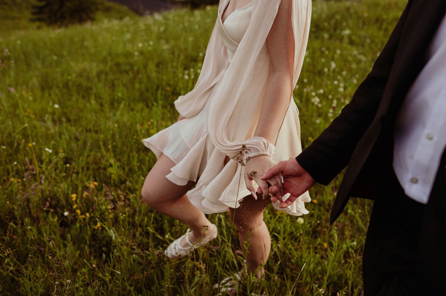 Close-up of a bride in a flowing cream dress and white sneakers holding hands with a groom in a meadow.