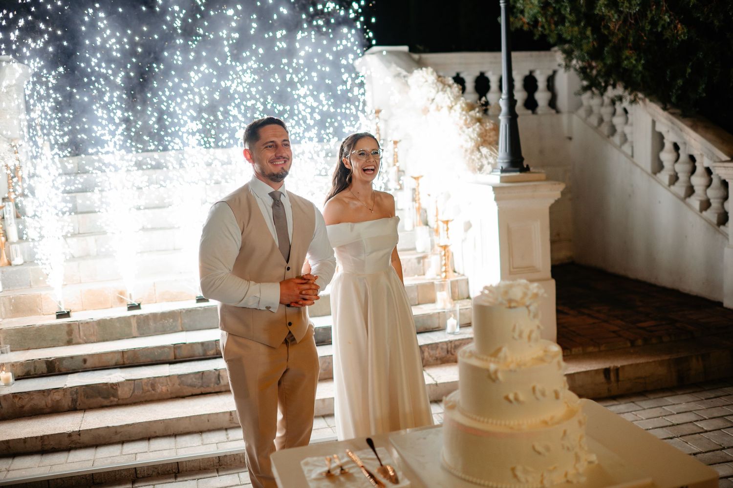 Smiling newlyweds pose beside white tiered wedding cake with dazzling light backdrop at night reception.