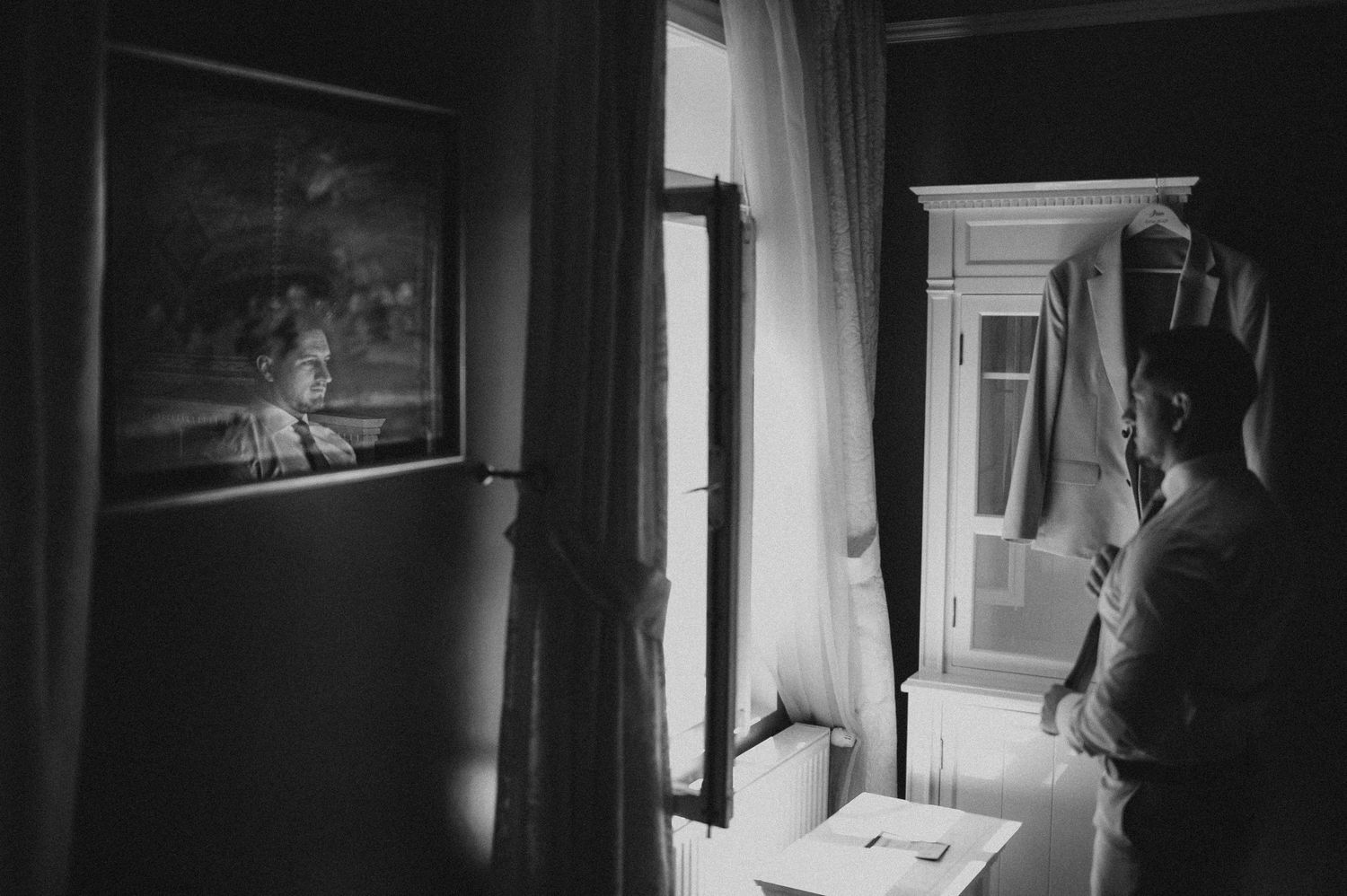 Groom preparing in dimly lit room by window in black and white photo.