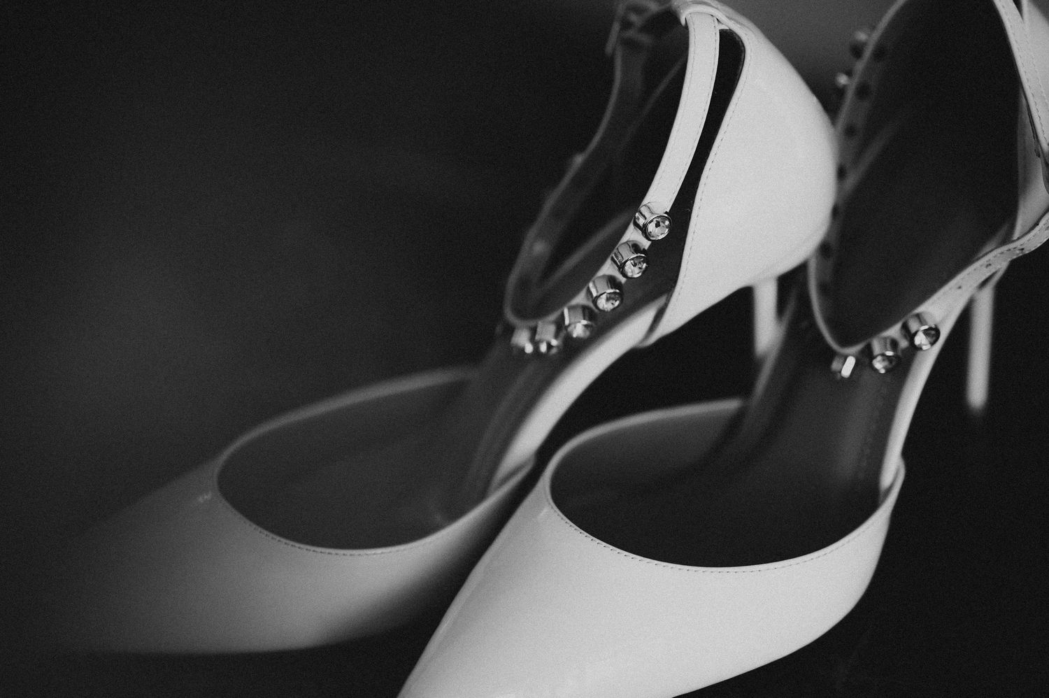 Black and white photo of bridal heels with crystal ankle straps on a dark background.