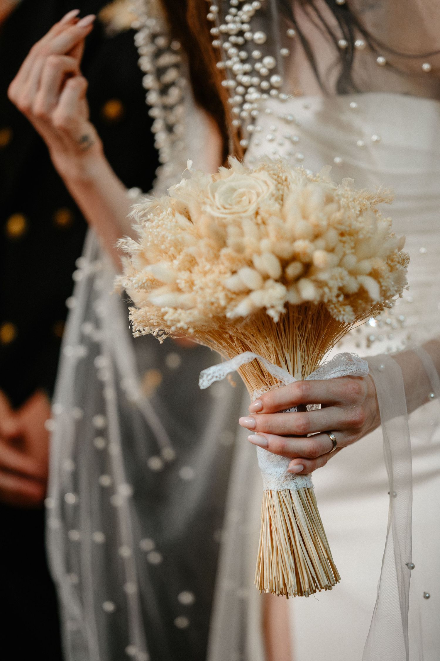 Close-up of bride holding elegant dried flower bouquet with pearl veil detail at wedding.