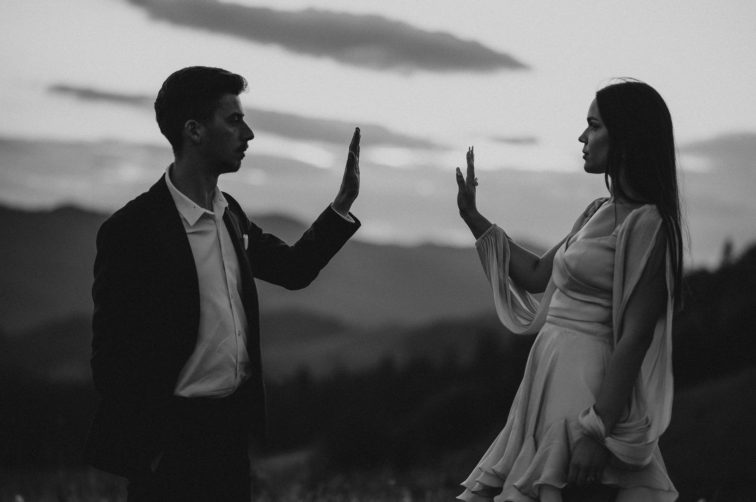 Black and white photo of a couple facing each other with raised hands outdoors at dusk.