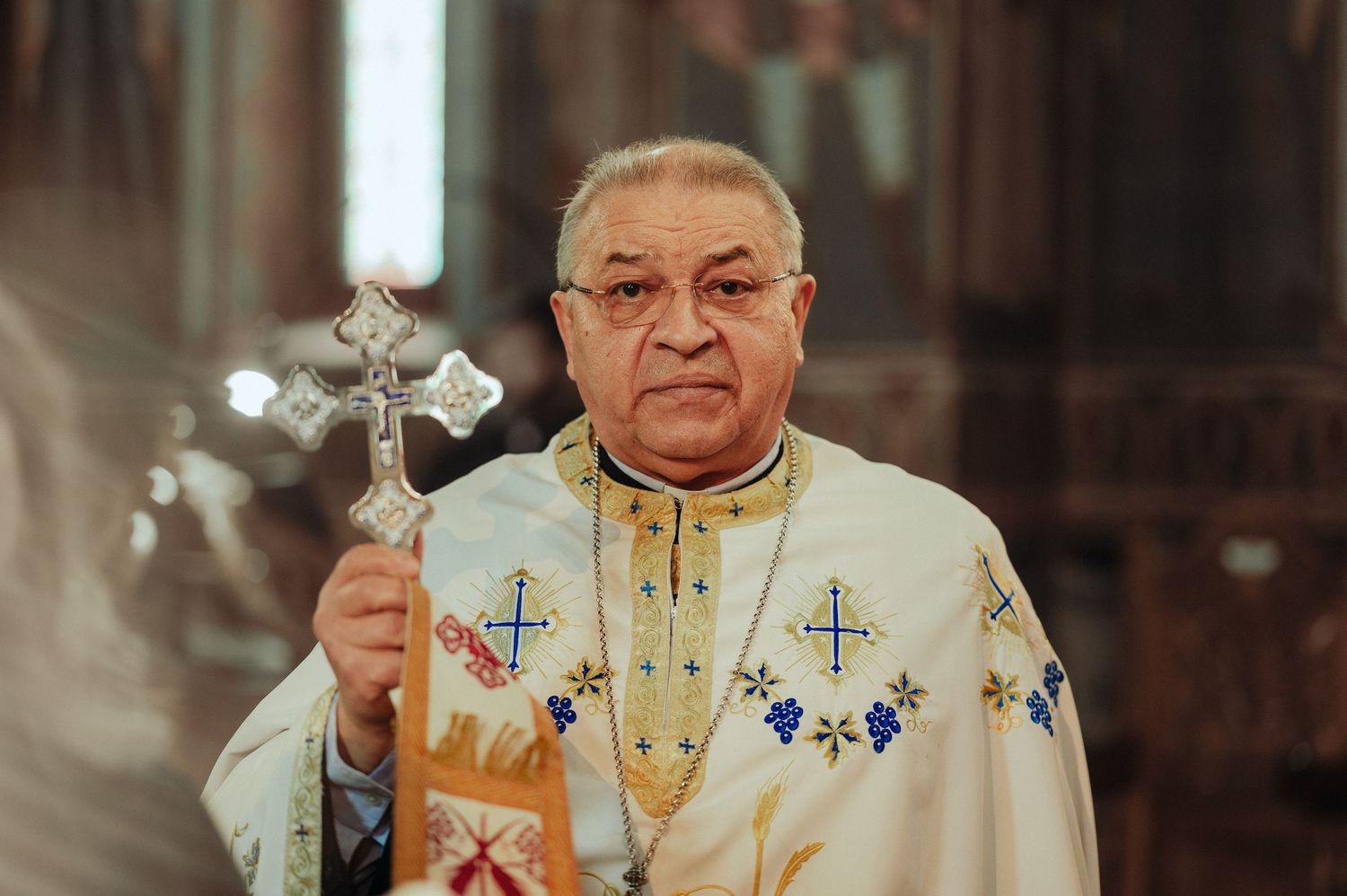 Orthodox priest in ornate white and gold vestments holding a silver cross during a ceremony.