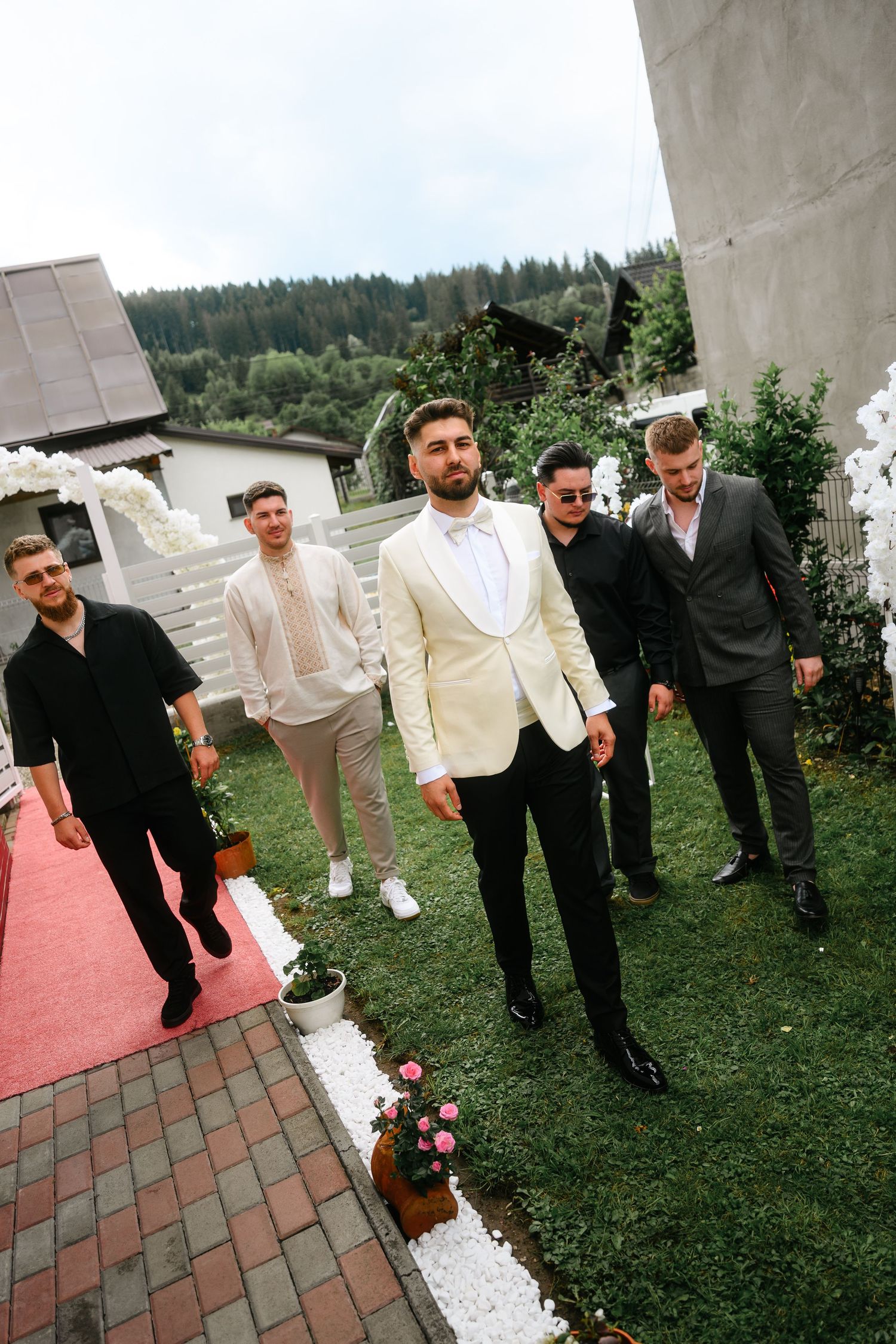 Groom in white tuxedo stands with groomsmen in black suits outdoors at a wedding venue with floral decor.