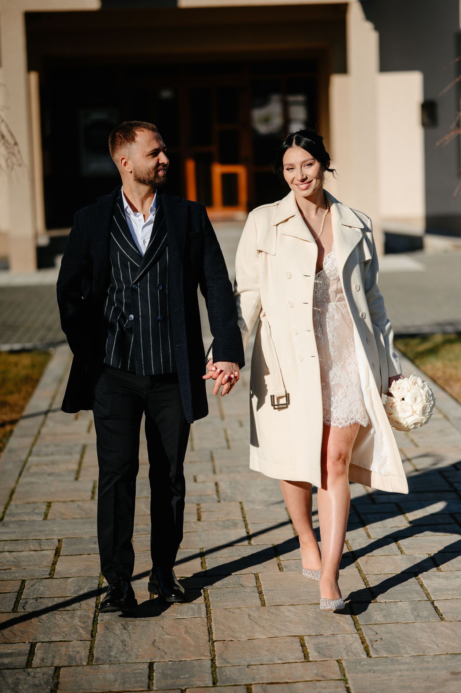 Elegant couple holding hands walking outdoors, bride in white lace dress and trench coat, groom in striped suit.
