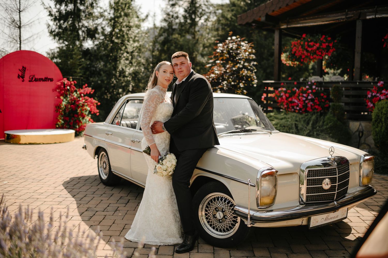 Bride and groom posing beside vintage white Mercedes-Benz at floral wedding venue entrance.