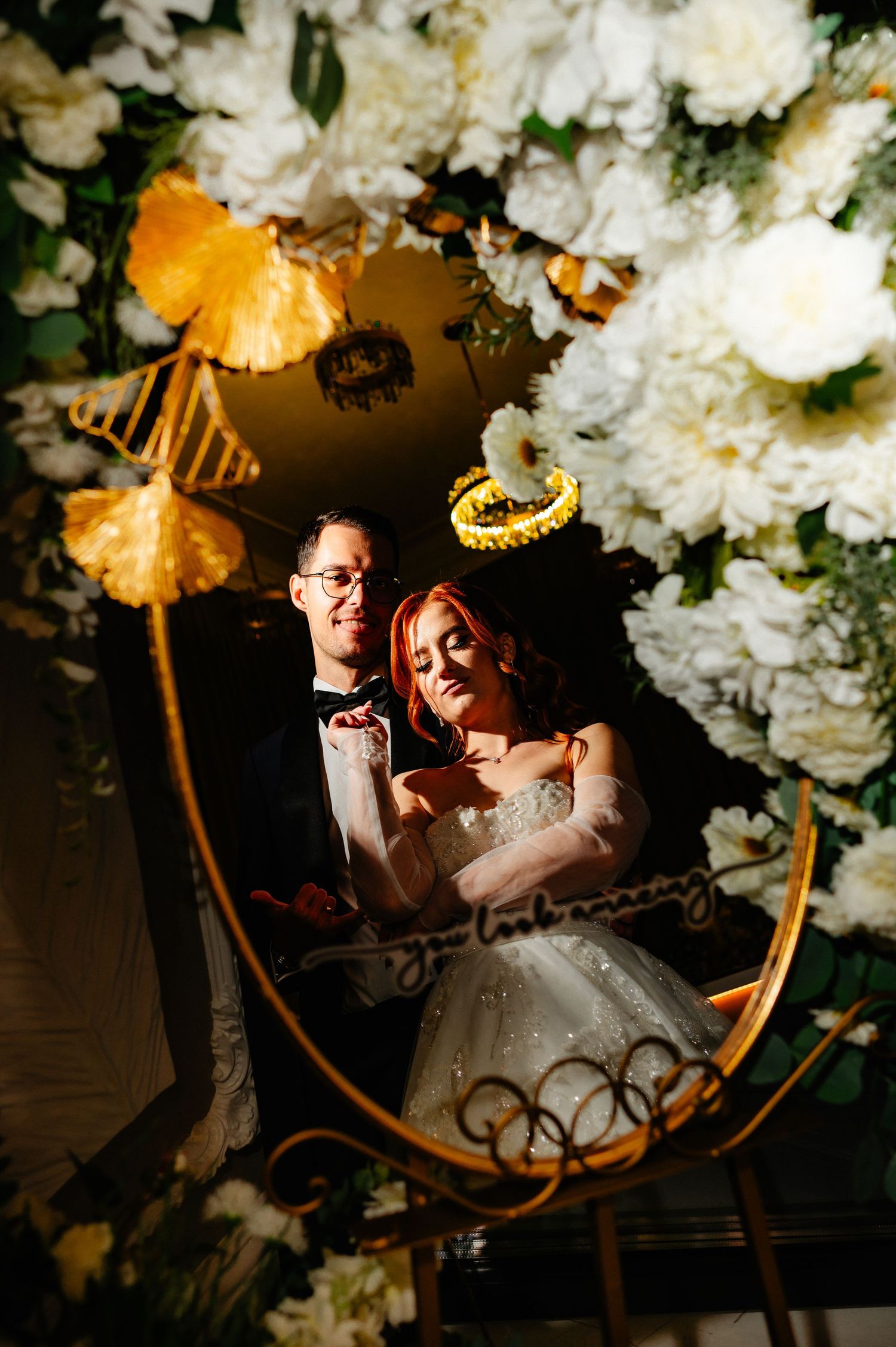 Bride and groom pose under a floral arch with white blooms and gold accents at an elegant wedding reception.