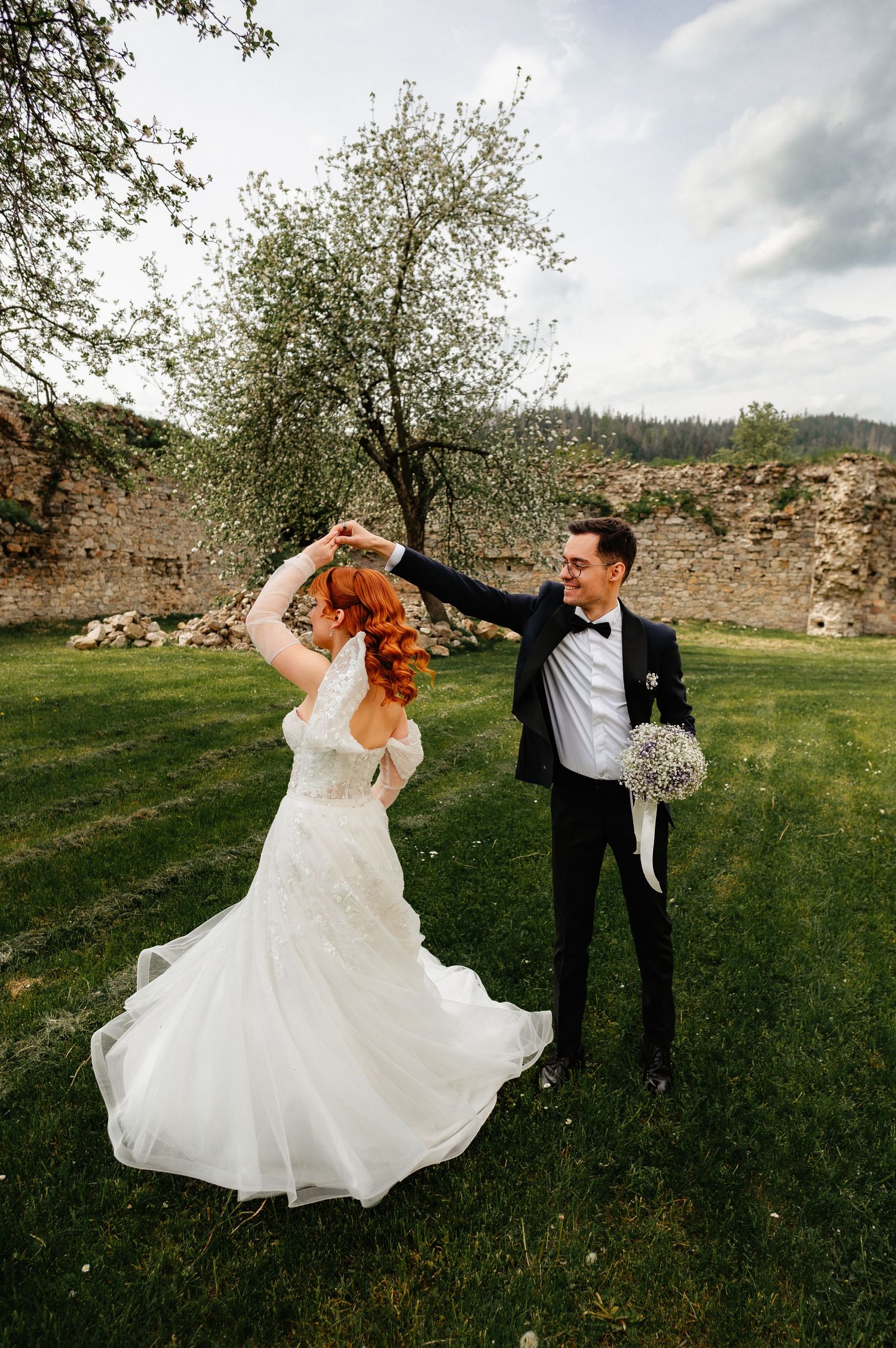 Groom in black tuxedo twirling red-haired bride in white wedding dress outdoors on green lawn near stone ruins.