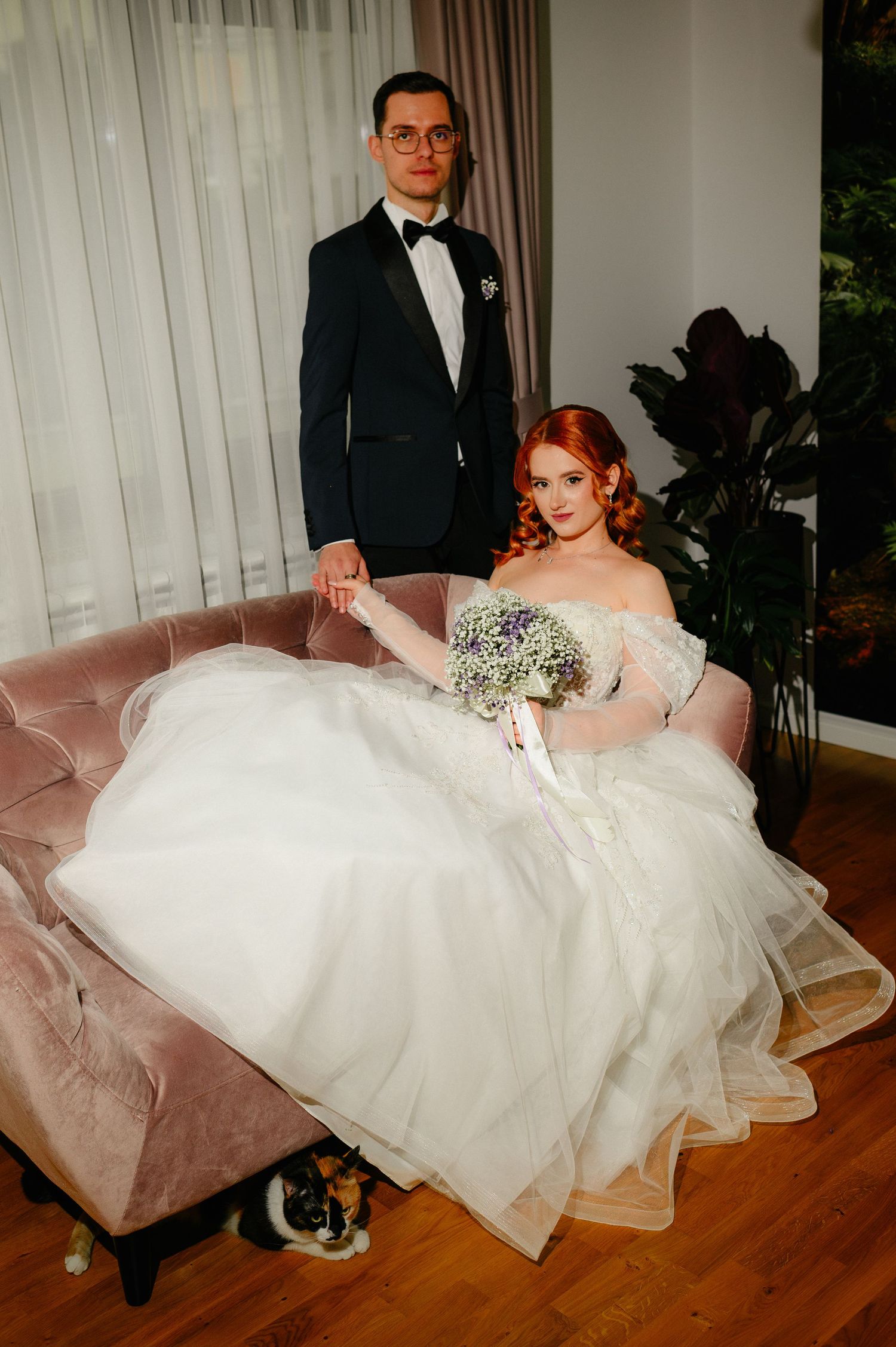 Bride in white gown sits on pink velvet sofa holding bouquet while groom in black tuxedo stands beside her.