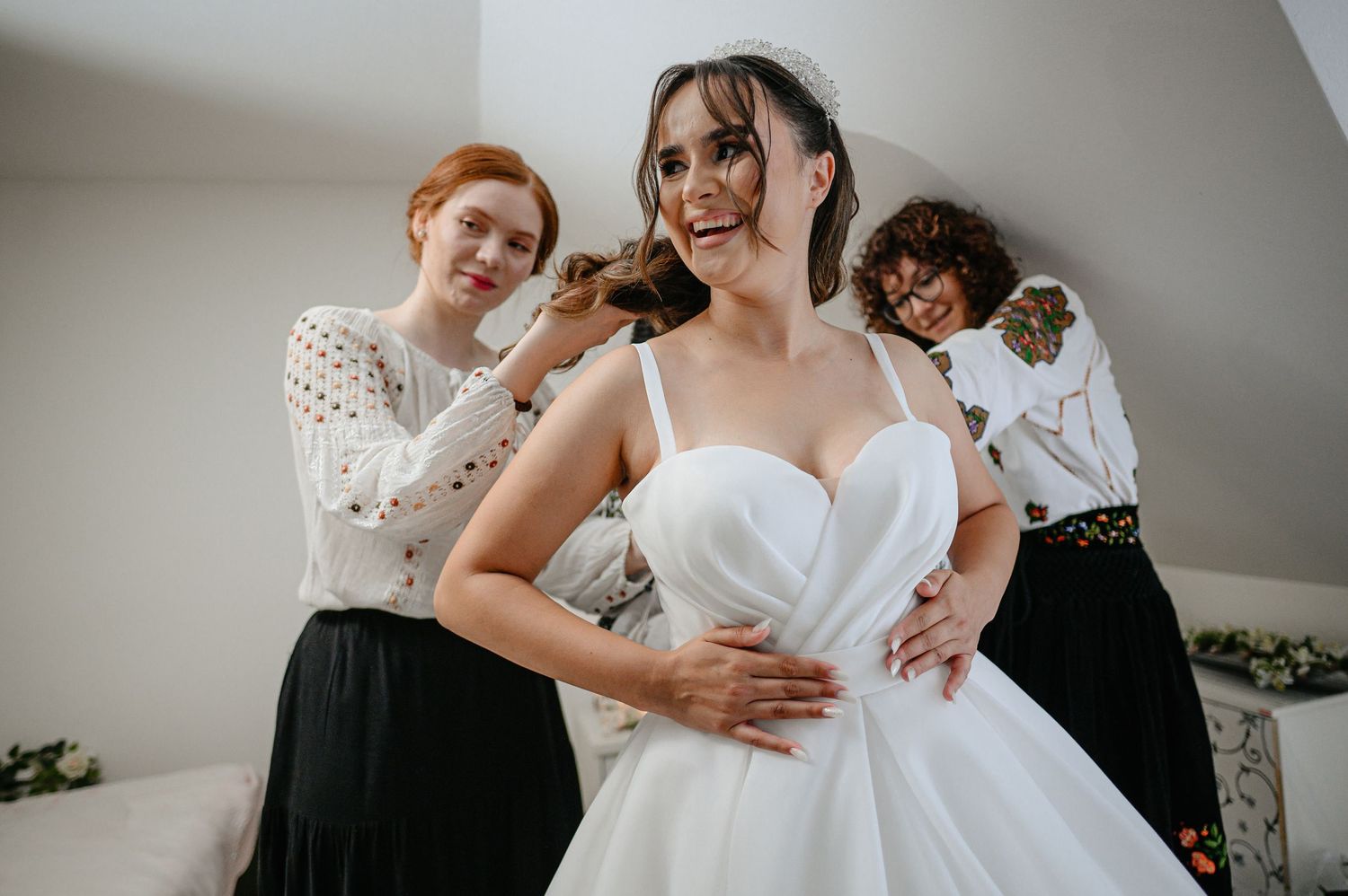 Two bridesmaids help a smiling bride into her white wedding gown before the ceremony.