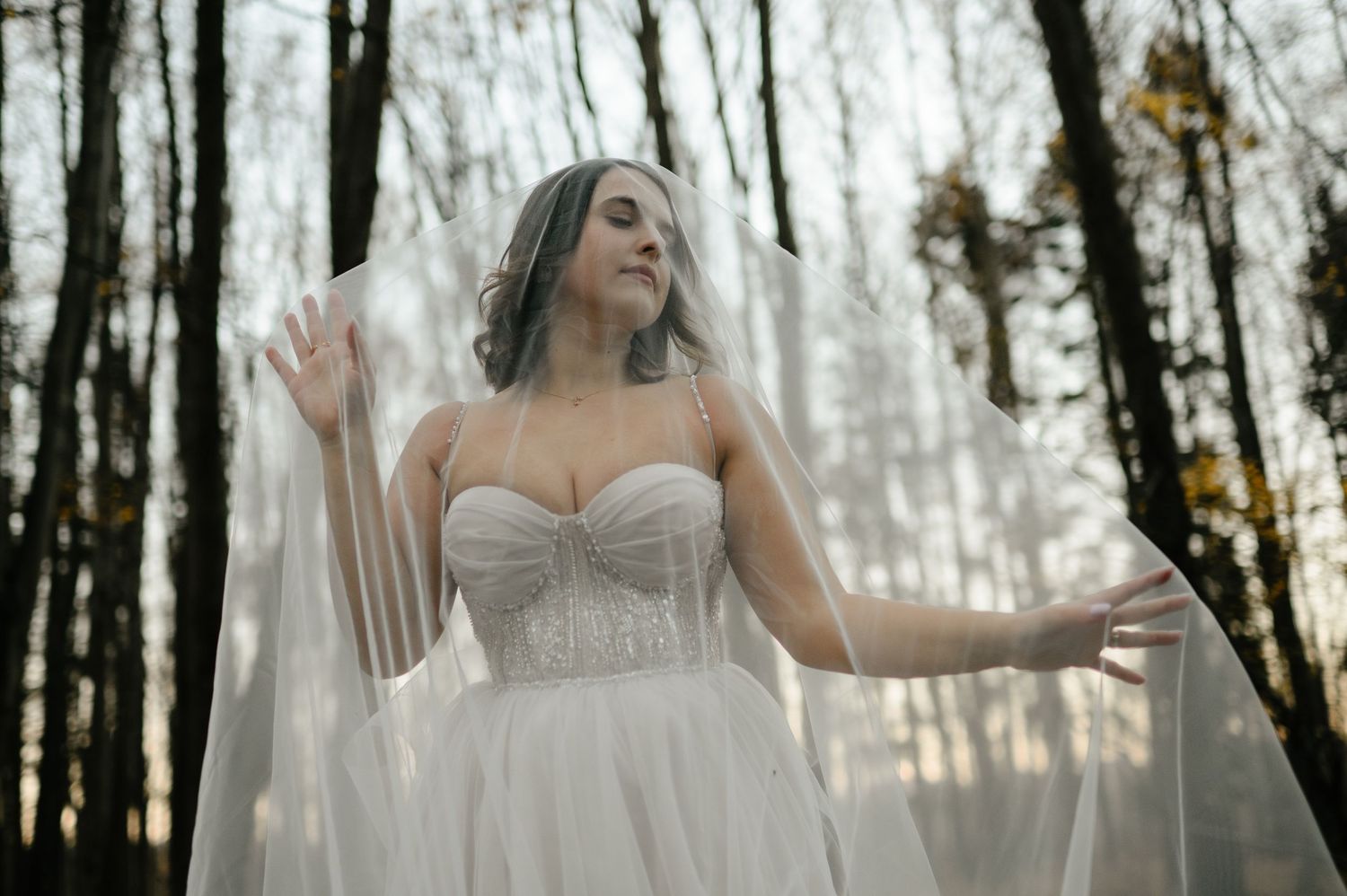 Bride in a strapless beaded wedding gown and flowing veil poses gracefully among tall trees in a forest setting.