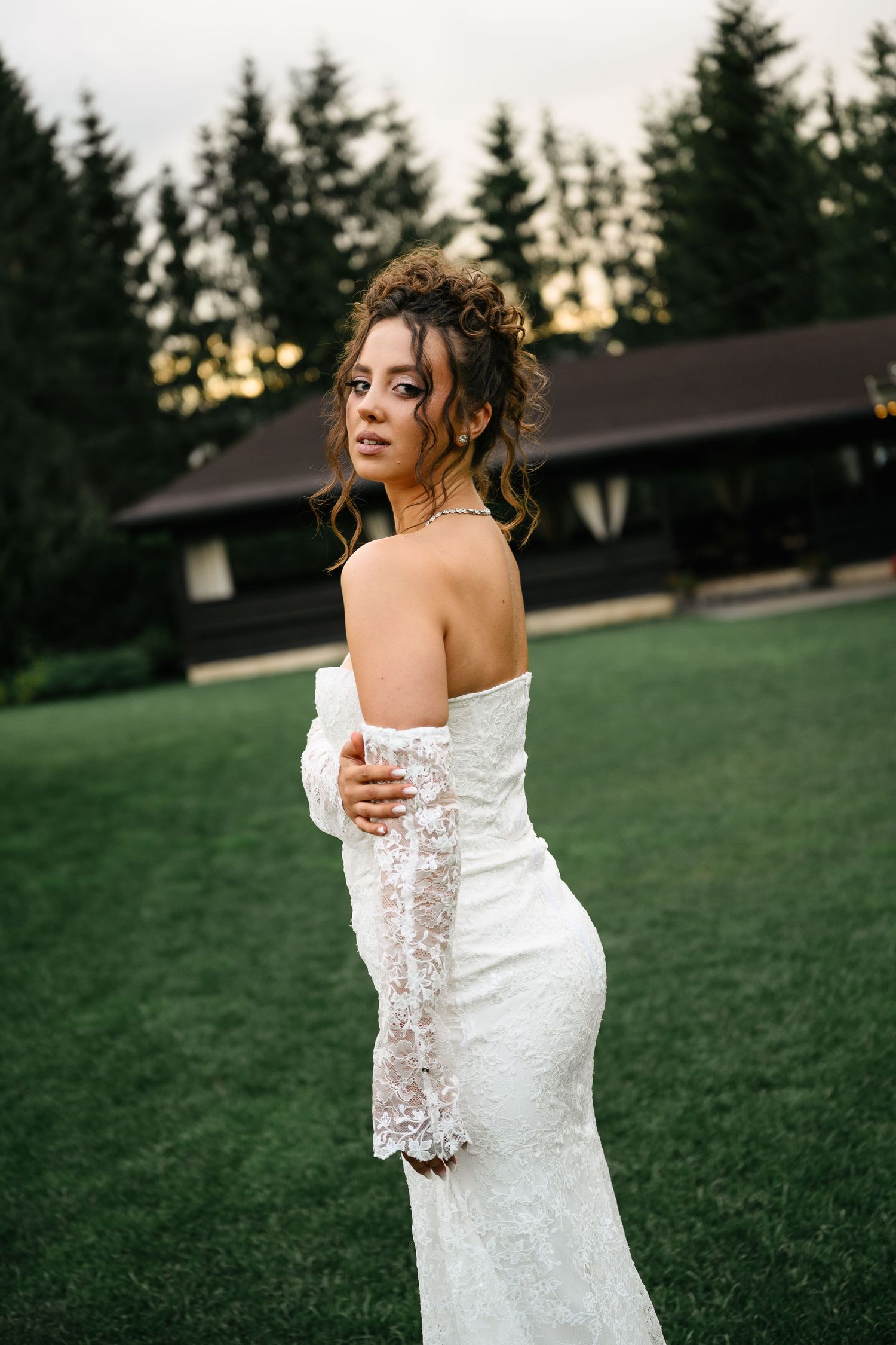 Elegant bride in white lace off-shoulder gown poses on green lawn with pine trees and rustic lodge backdrop.