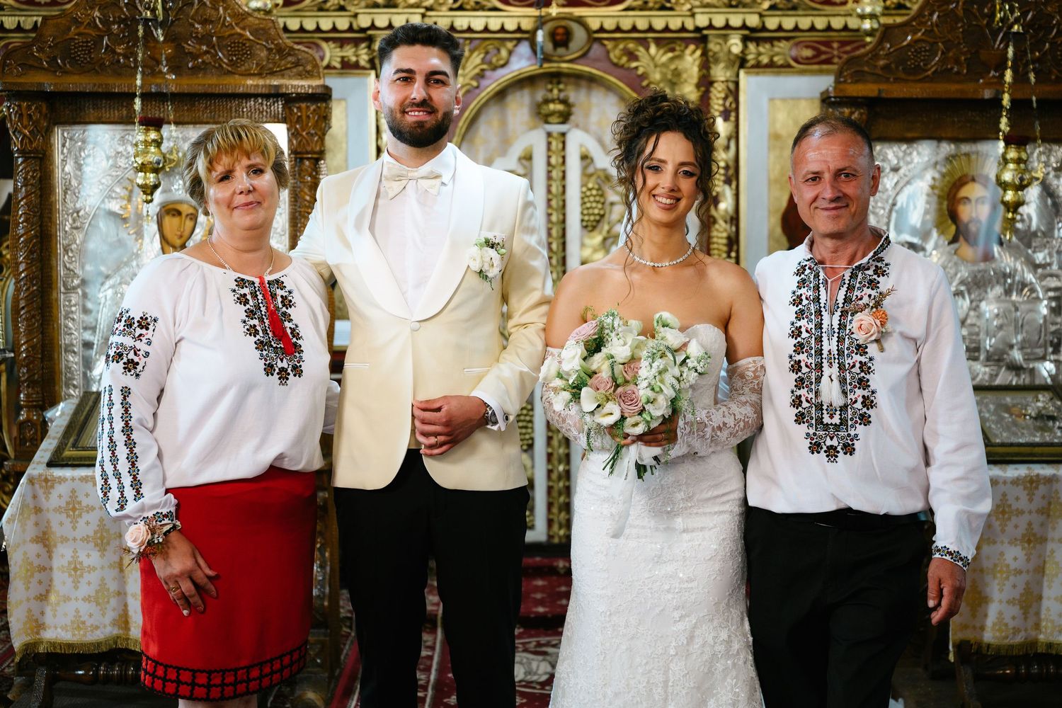 Bride and groom pose with family in traditional Romanian embroidered attire inside an ornate Orthodox church.