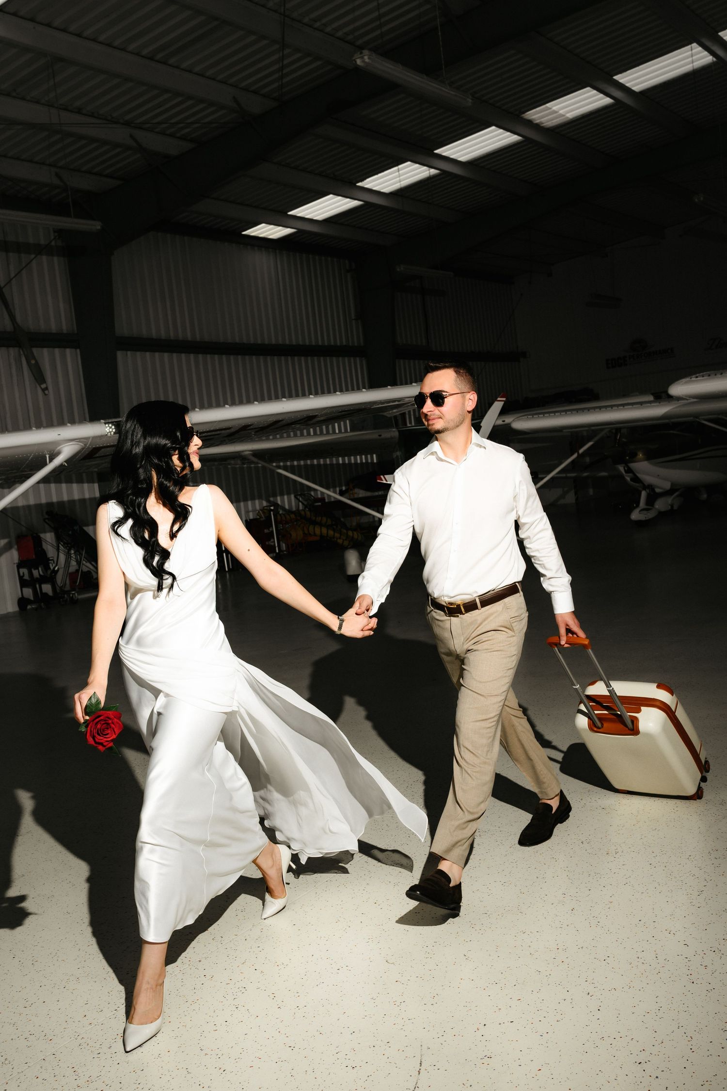 Bride in white dress and groom in sunglasses hold hands walking through aircraft hangar with luggage.