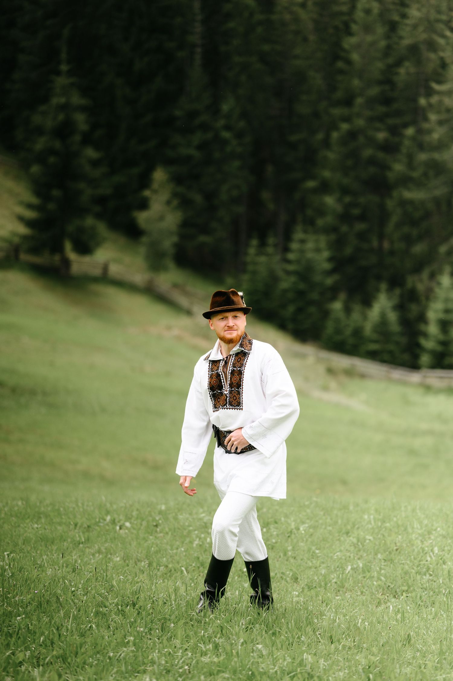 Man in traditional Eastern European folk costume with white shirt, embroidery, hat, and boots standing in green meadow.