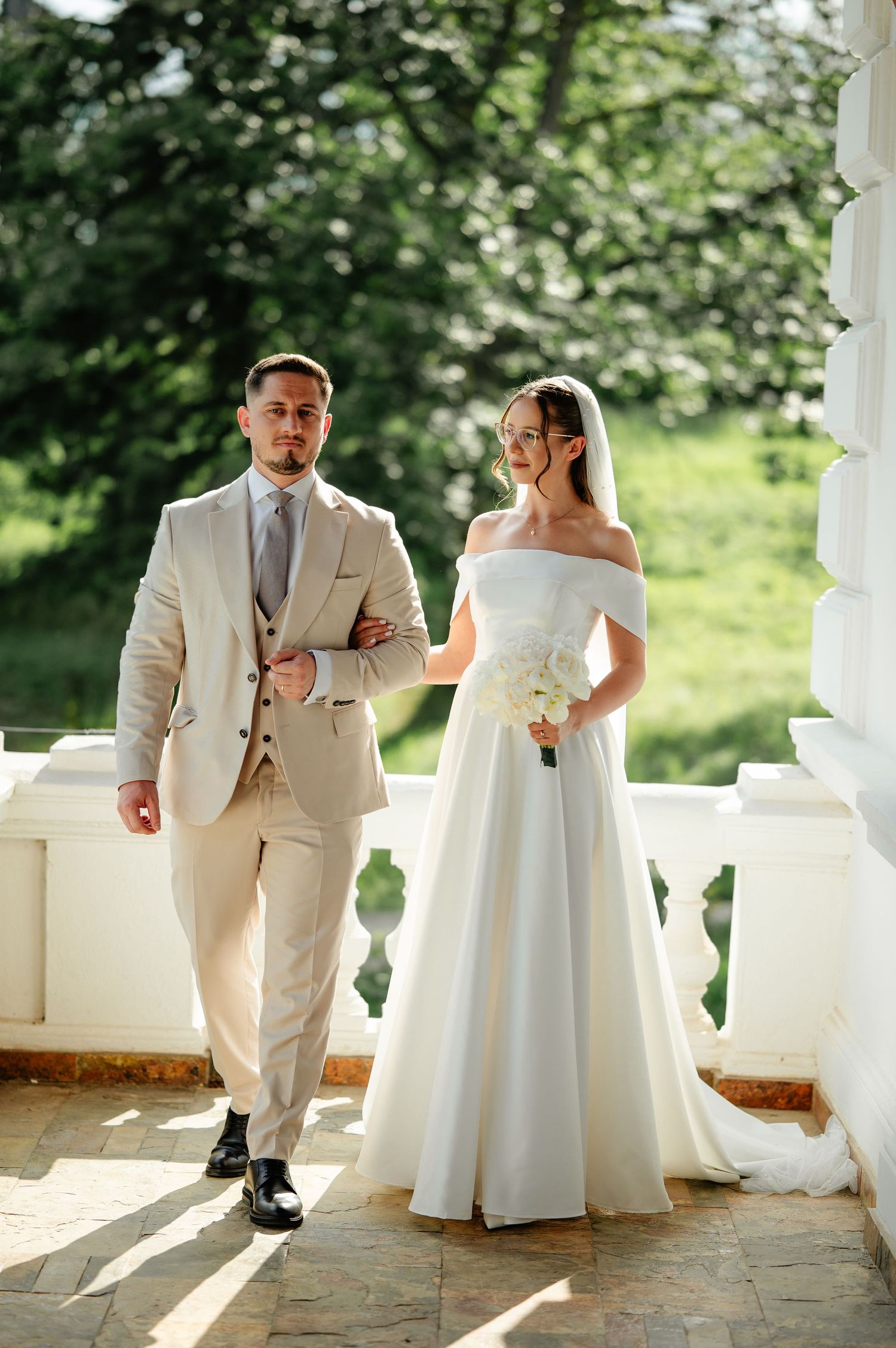 Elegant bride in off-shoulder white gown with veil walks arm-in-arm with groom in beige suit on sunlit balcony.