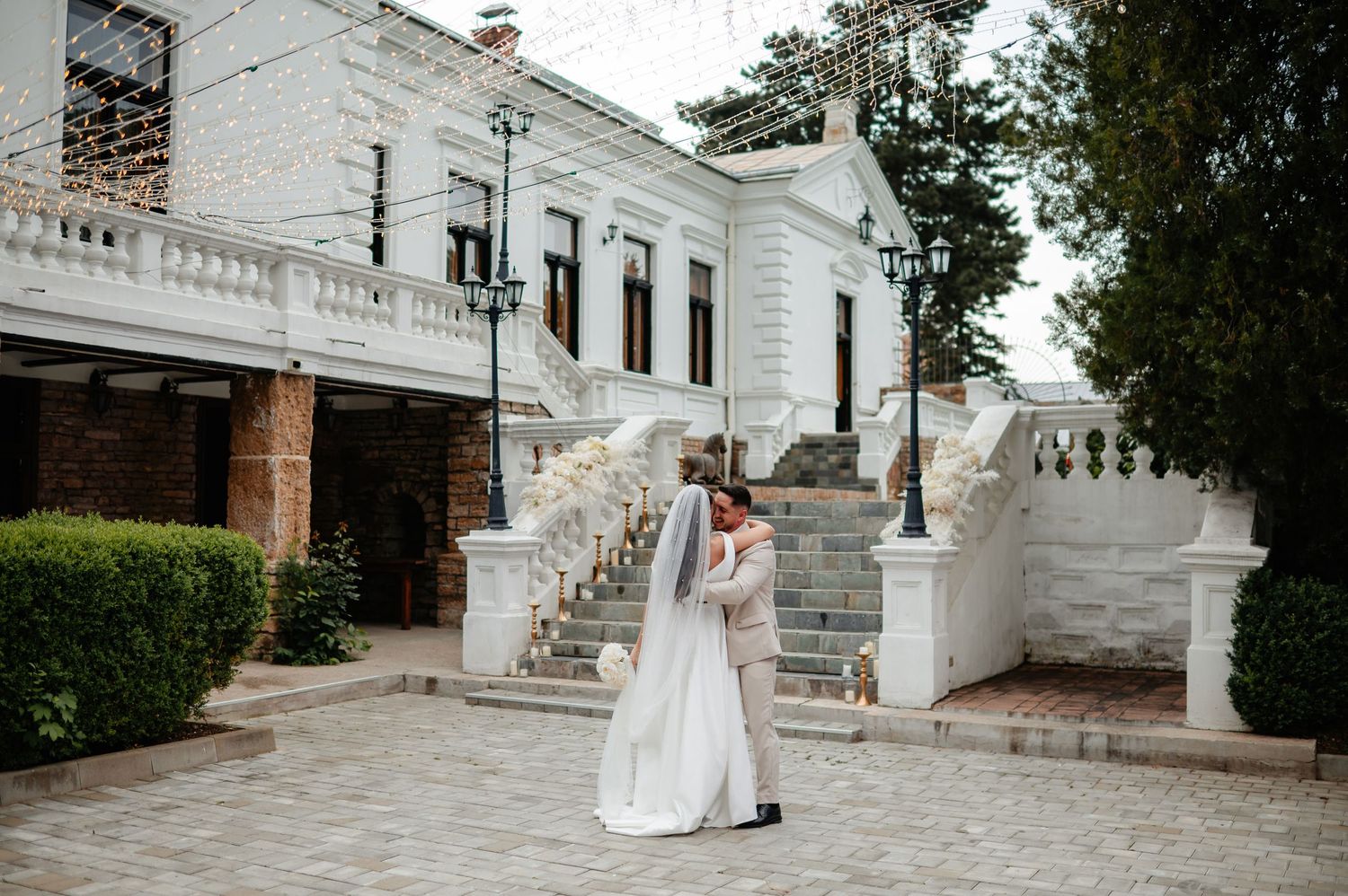 Bride and groom share romantic kiss in courtyard of white historic wedding venue with grand staircase and lamp posts.
