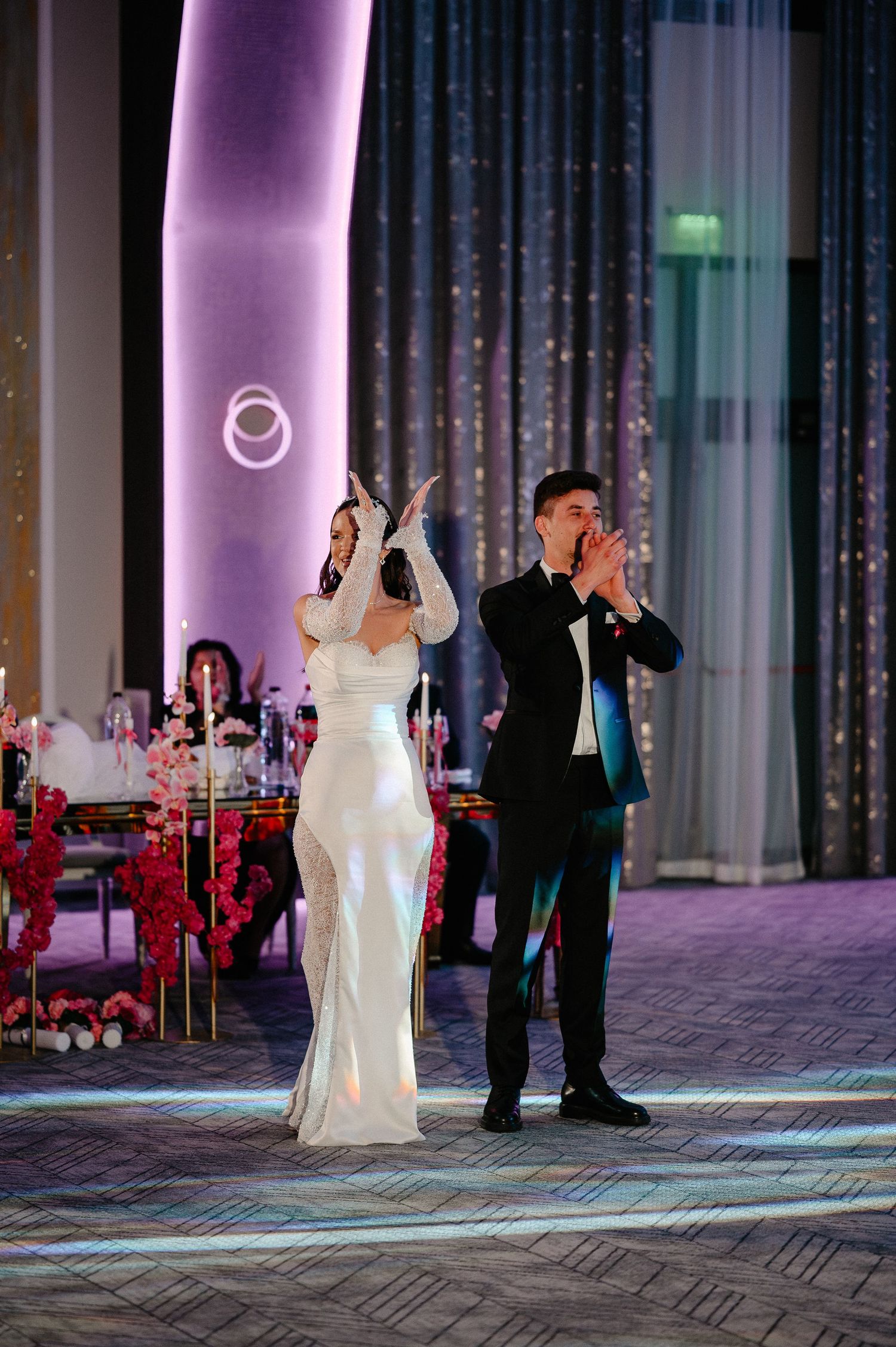 Bride in white gown and groom in black tuxedo clapping during their first dance at elegant wedding reception.